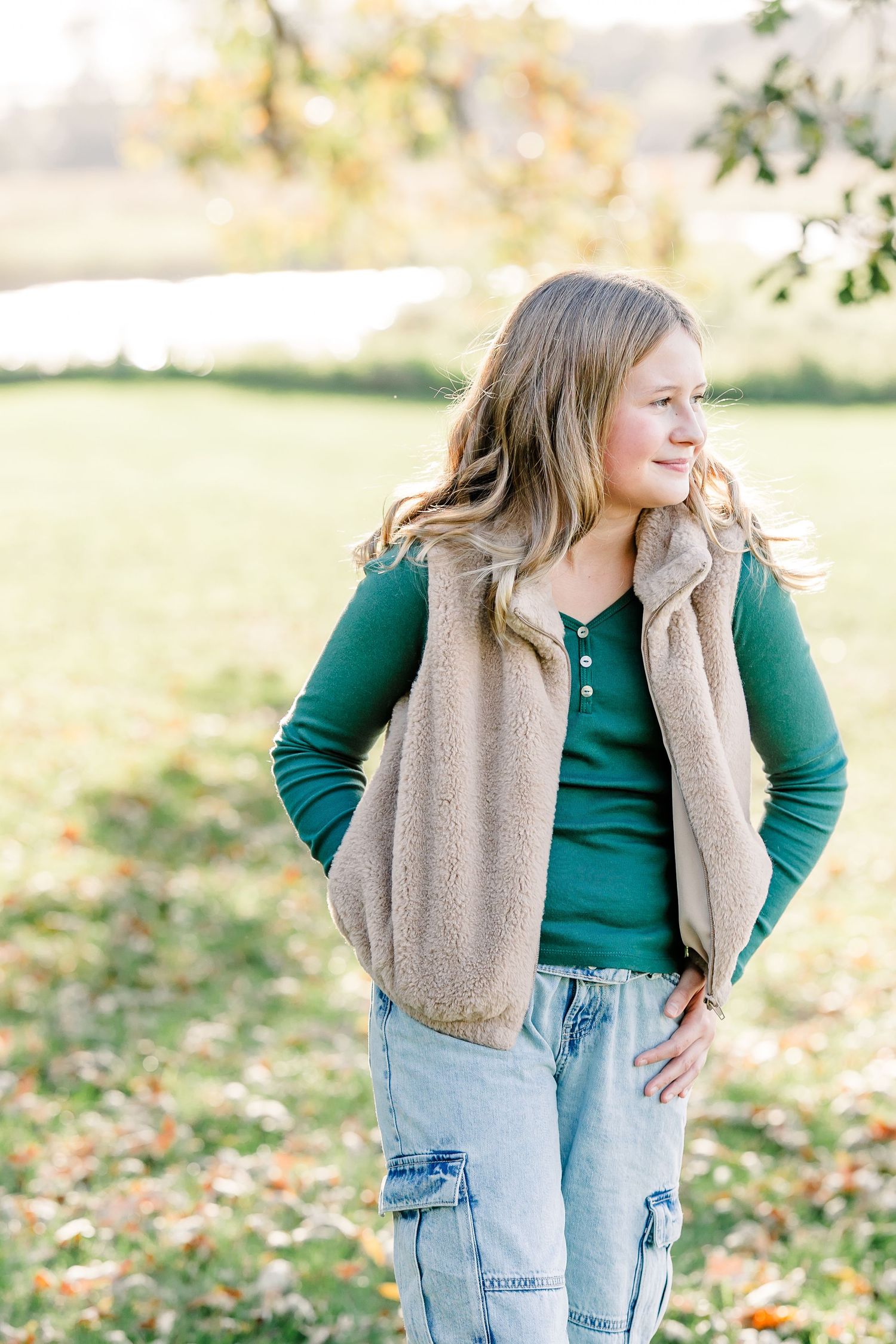 Young girl in a green top, tan fuzzy vest and cargo jeans stands smiling with hands on hips on a leaf-covered lawn.