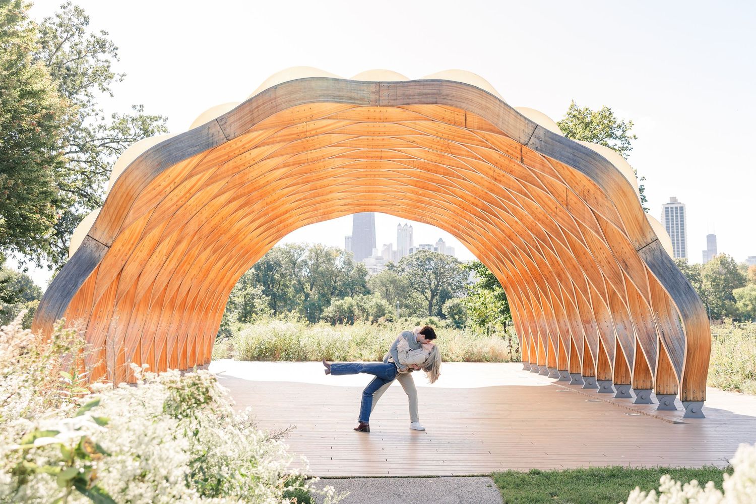Couple dancing under an orange wooden arch pavilion in a sunny park with city skyline in the background.