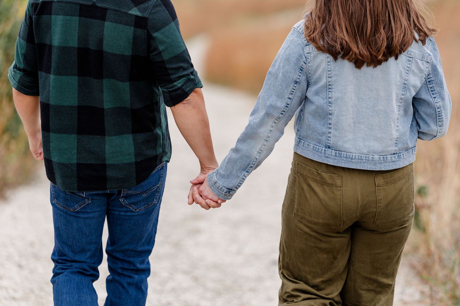 Couple holding hands while walking down a gravel path, shown from behind, he in a plaid shirt and she in a denim jacket.