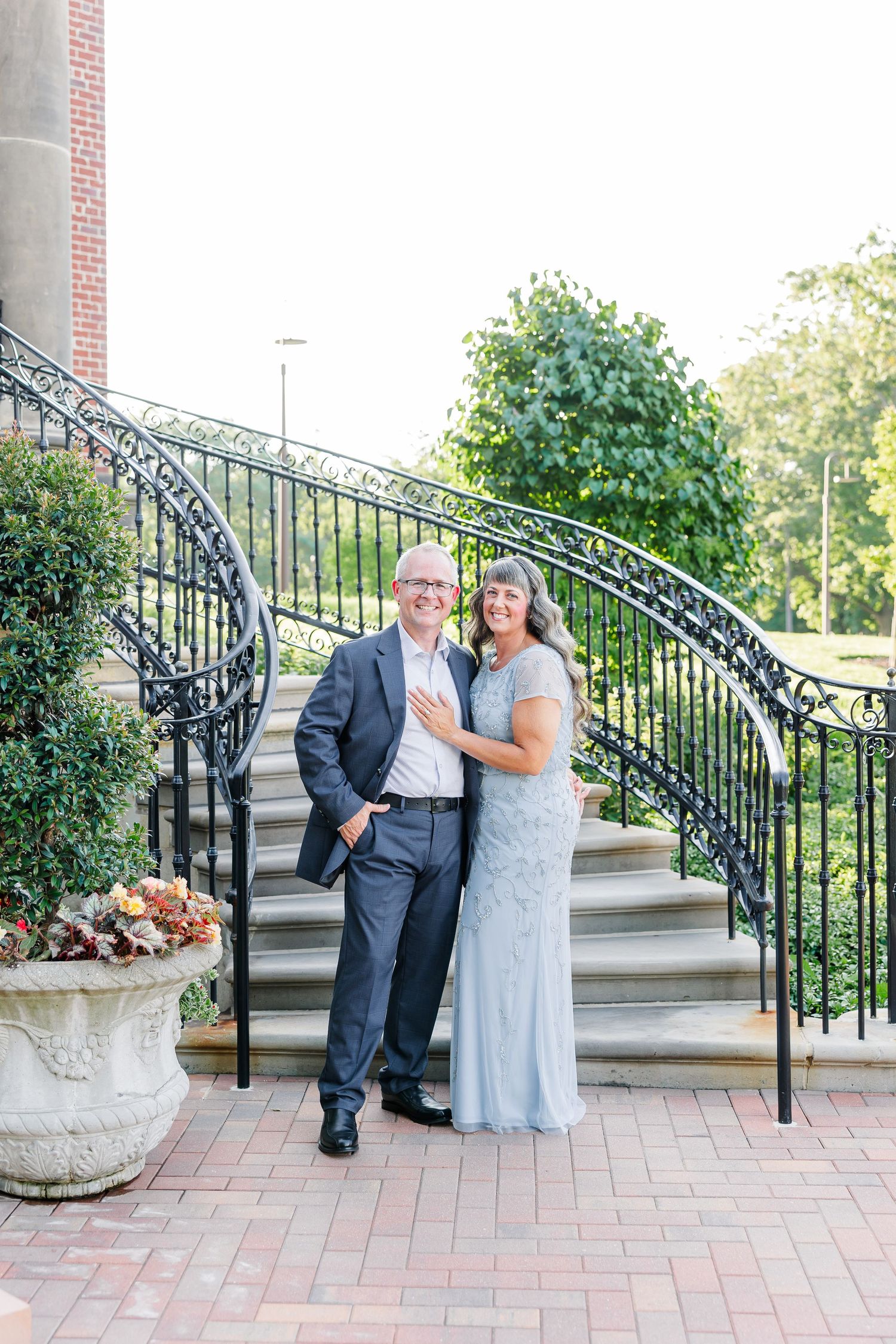 Smiling couple in formal attire poses on brick steps with ornate iron railing and lush greenery backdrop.