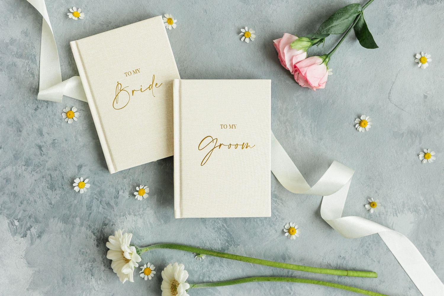 Gold foil To My Bride and To My Groom wedding journals surrounded by flowers and ribbon on gray background.
