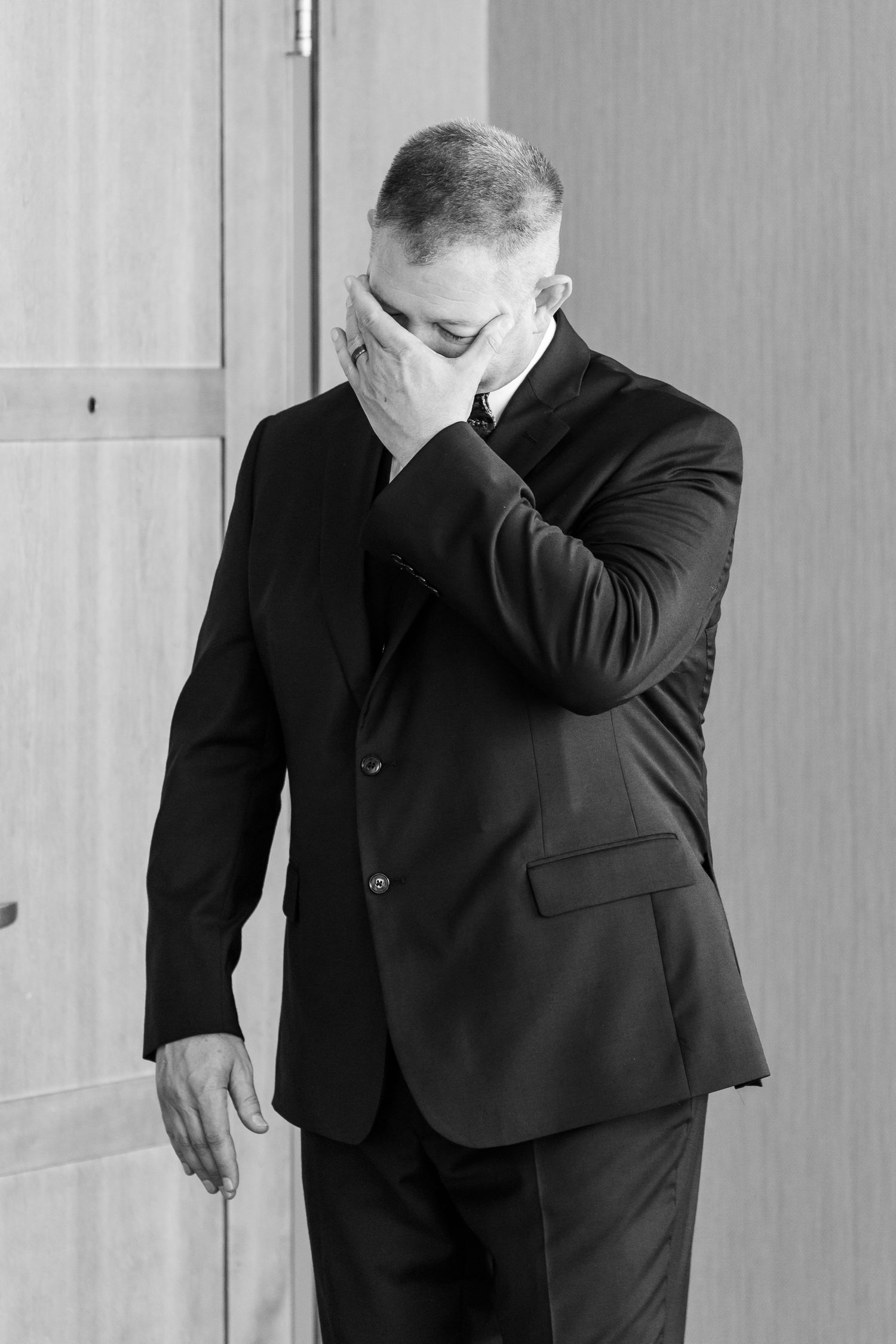 Emotional groom in black suit covers face with hand during wedding day first look moment.