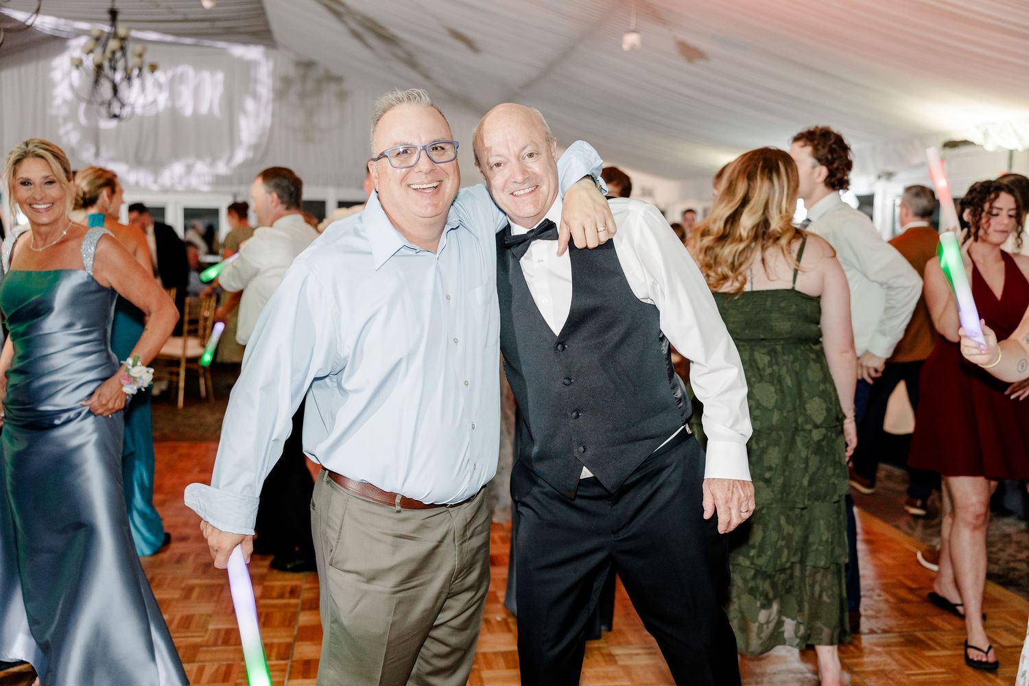 Two smiling men posing together with glow sticks on lively wedding reception dance floor.