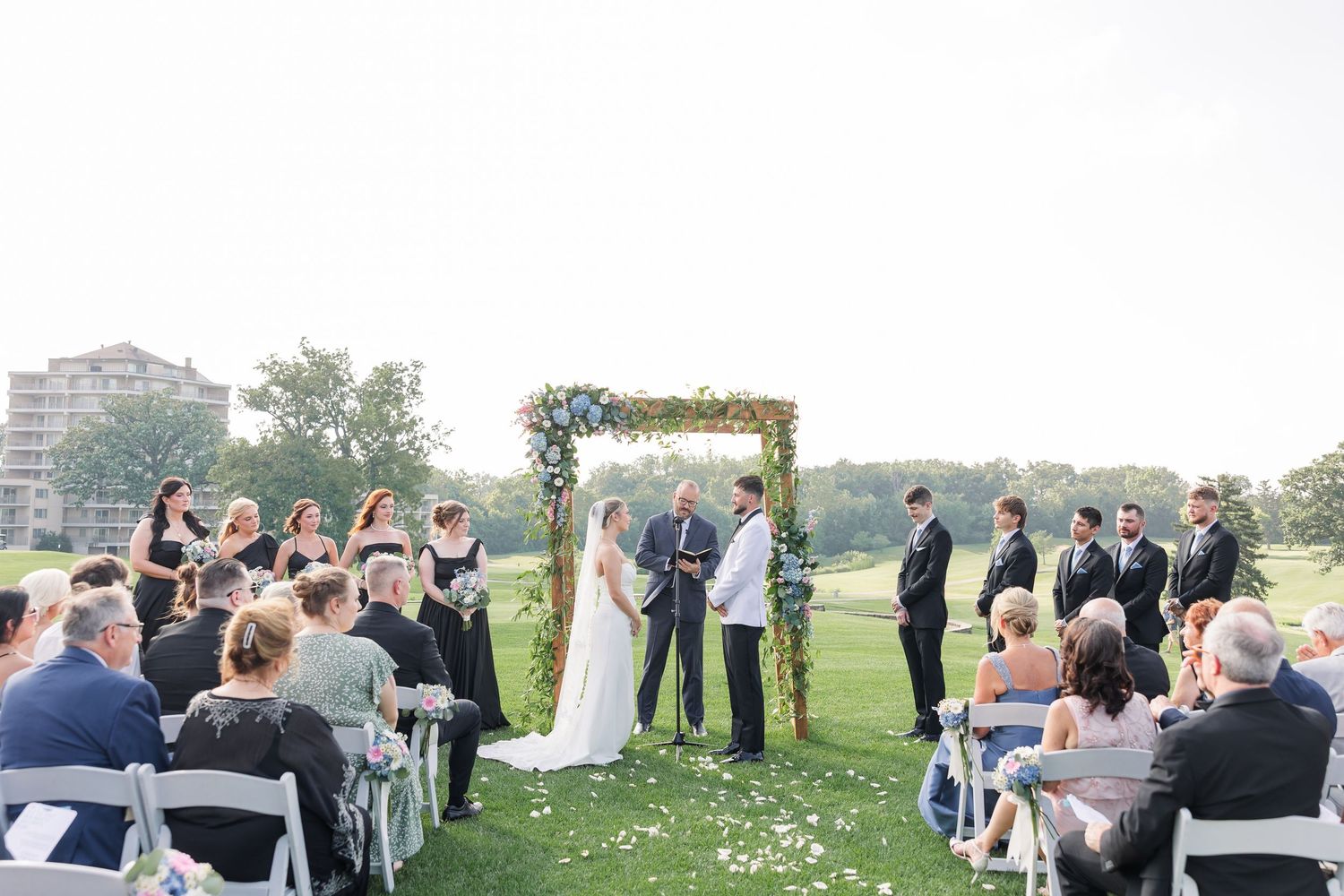 Bride and groom exchange vows at floral arch altar on golf course with seated wedding guests.