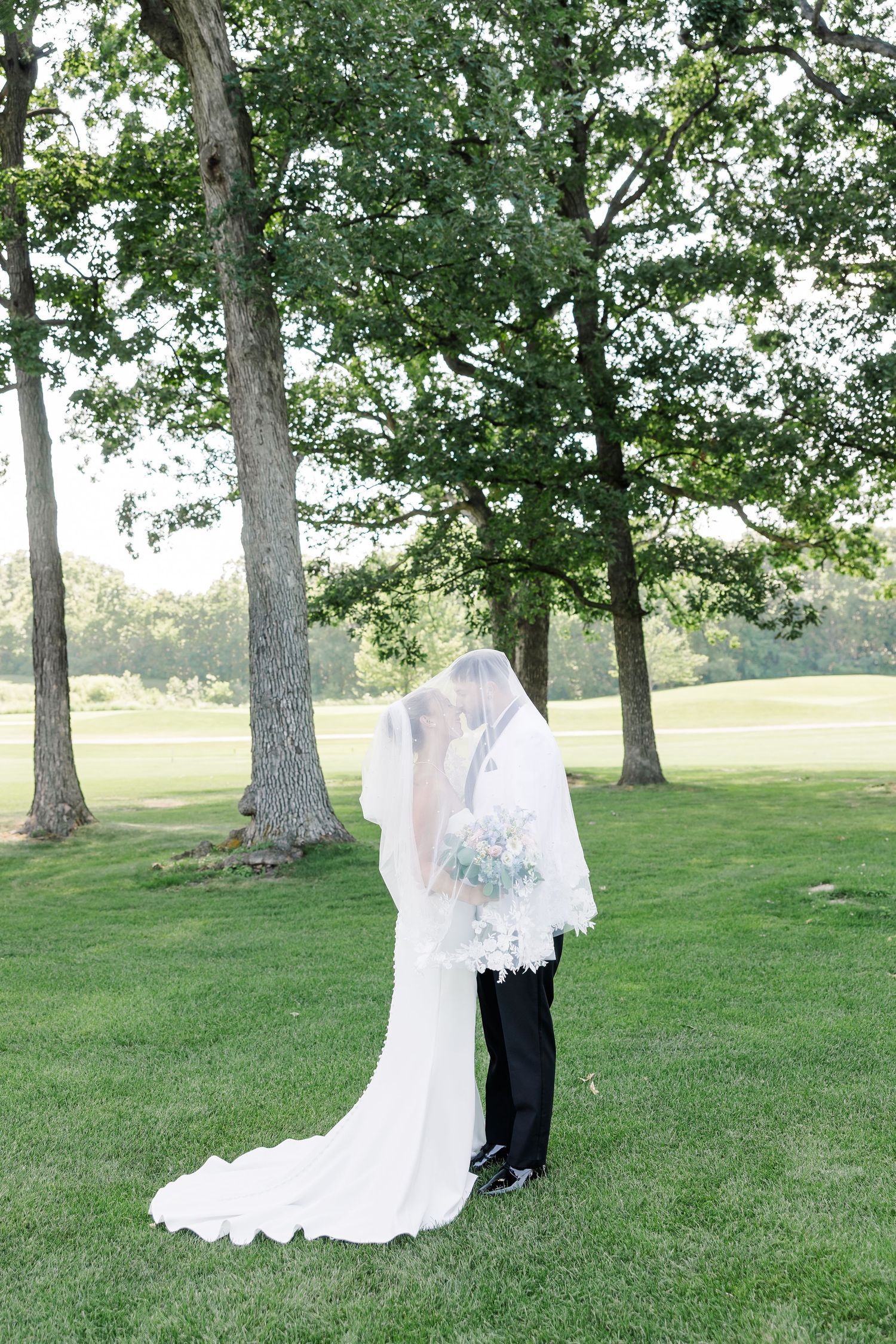 Bride and groom sharing a kiss under a flowing lace veil on a lush green lawn surrounded by tall oak trees.