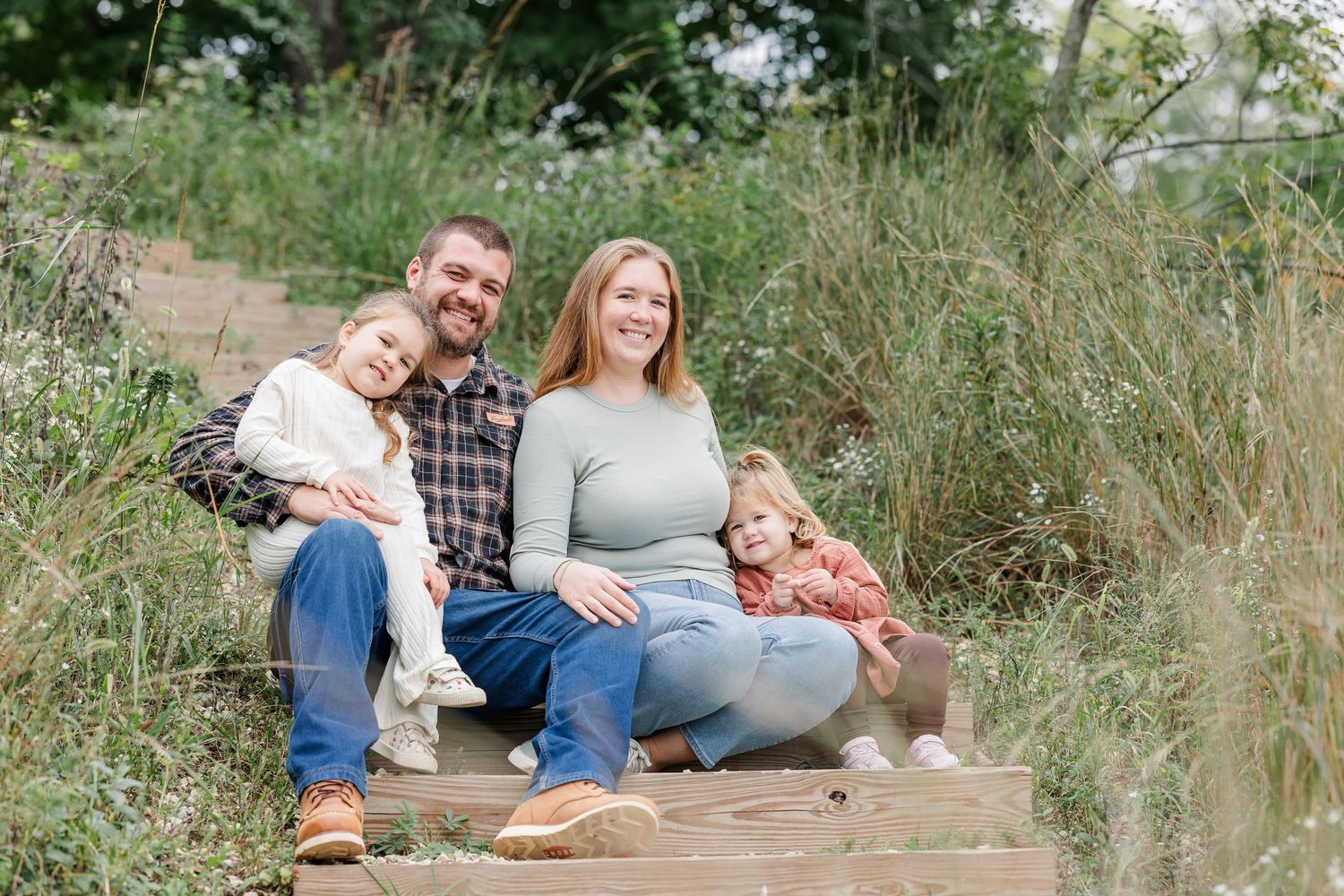 Happy family of four sitting on wooden steps surrounded by tall green grass during an outdoor fall photo session.