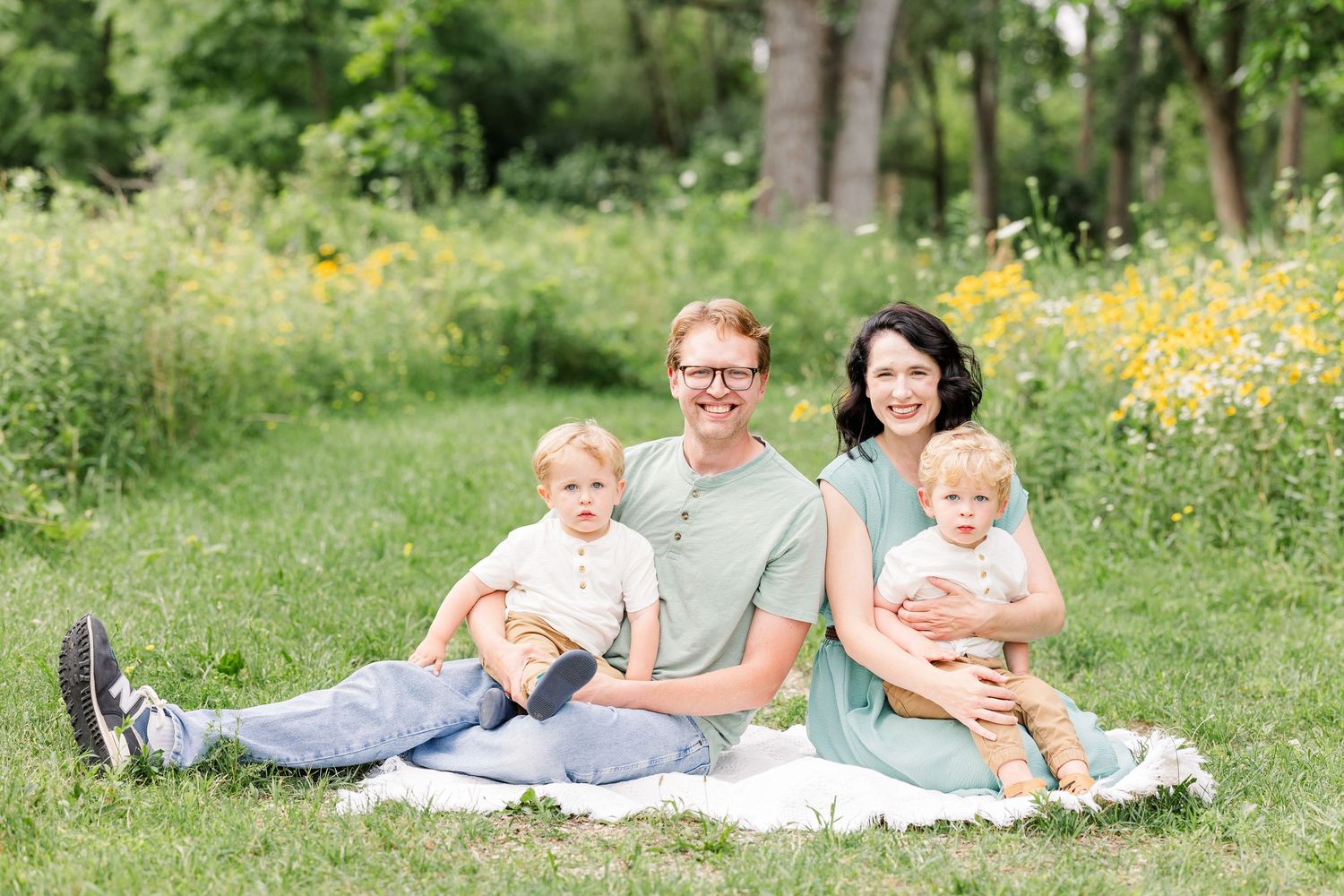Family of four smiling on a white blanket in a sunny green meadow with yellow wildflowers in the background.