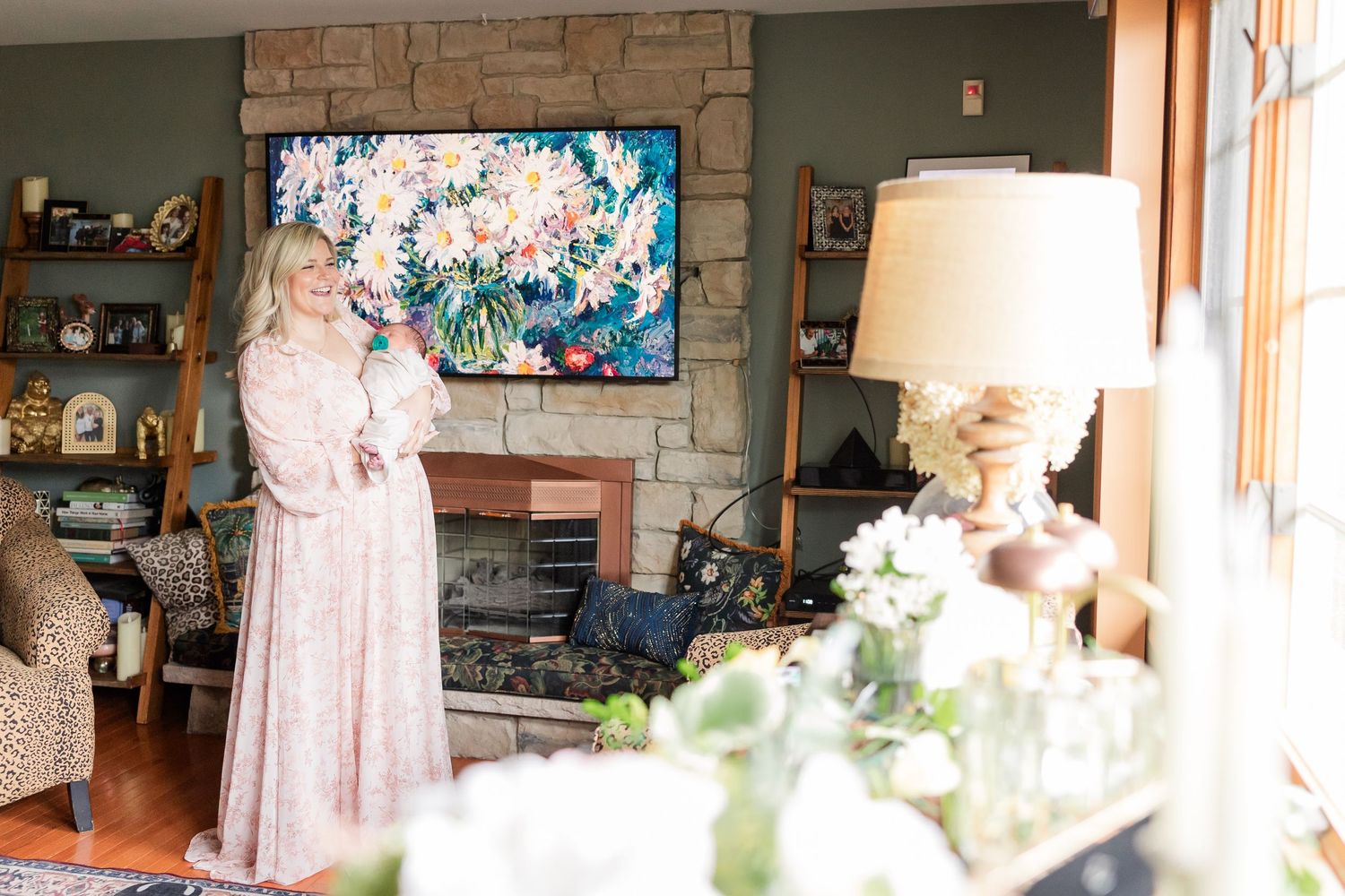 Woman in pink dress stands by stone fireplace with floral artwork above it in an elegantly decorated living room.