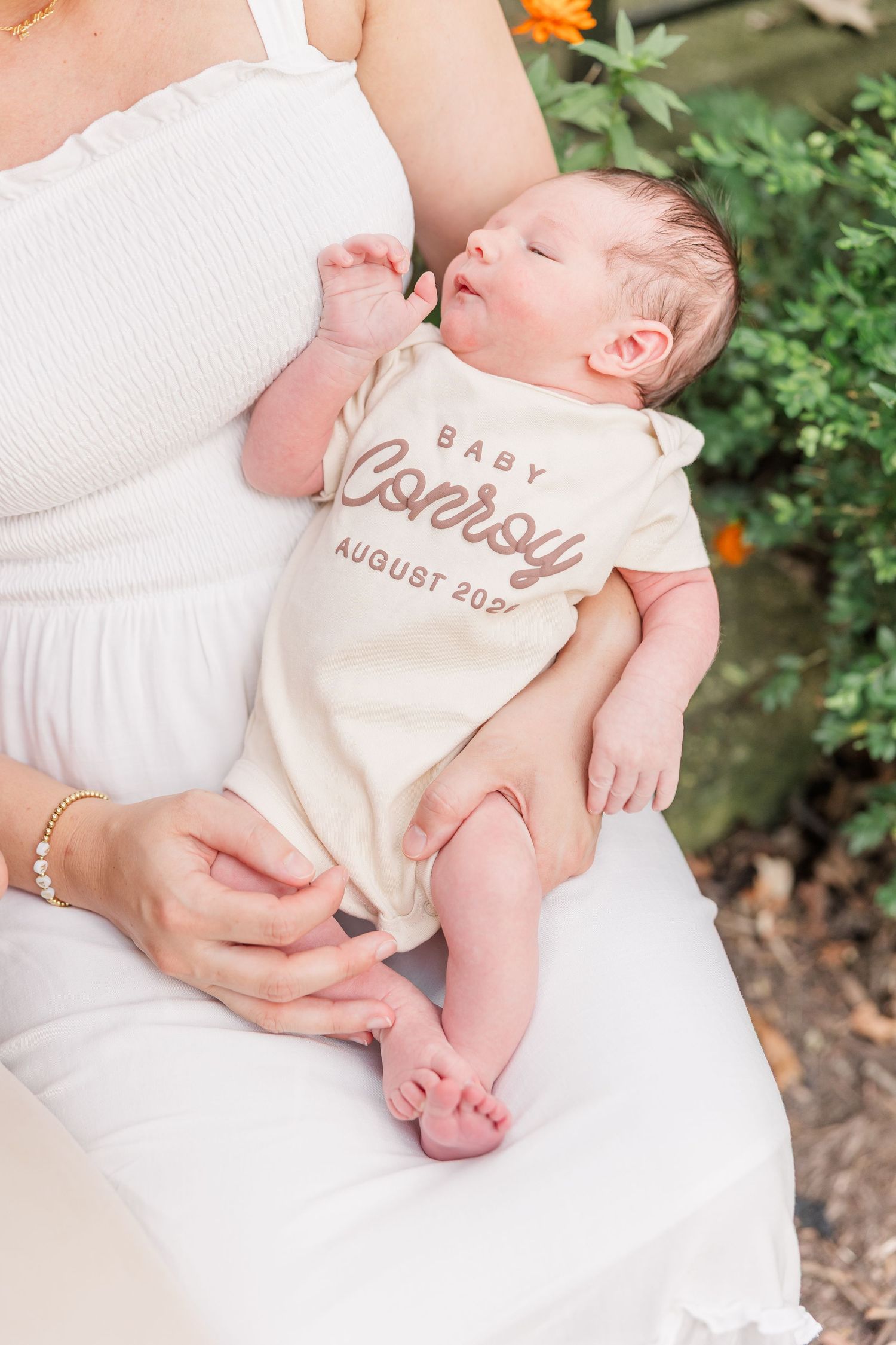 Mother in white dress holding newborn baby wearing a personalized cream romper reading Baby Conroy August 2024.