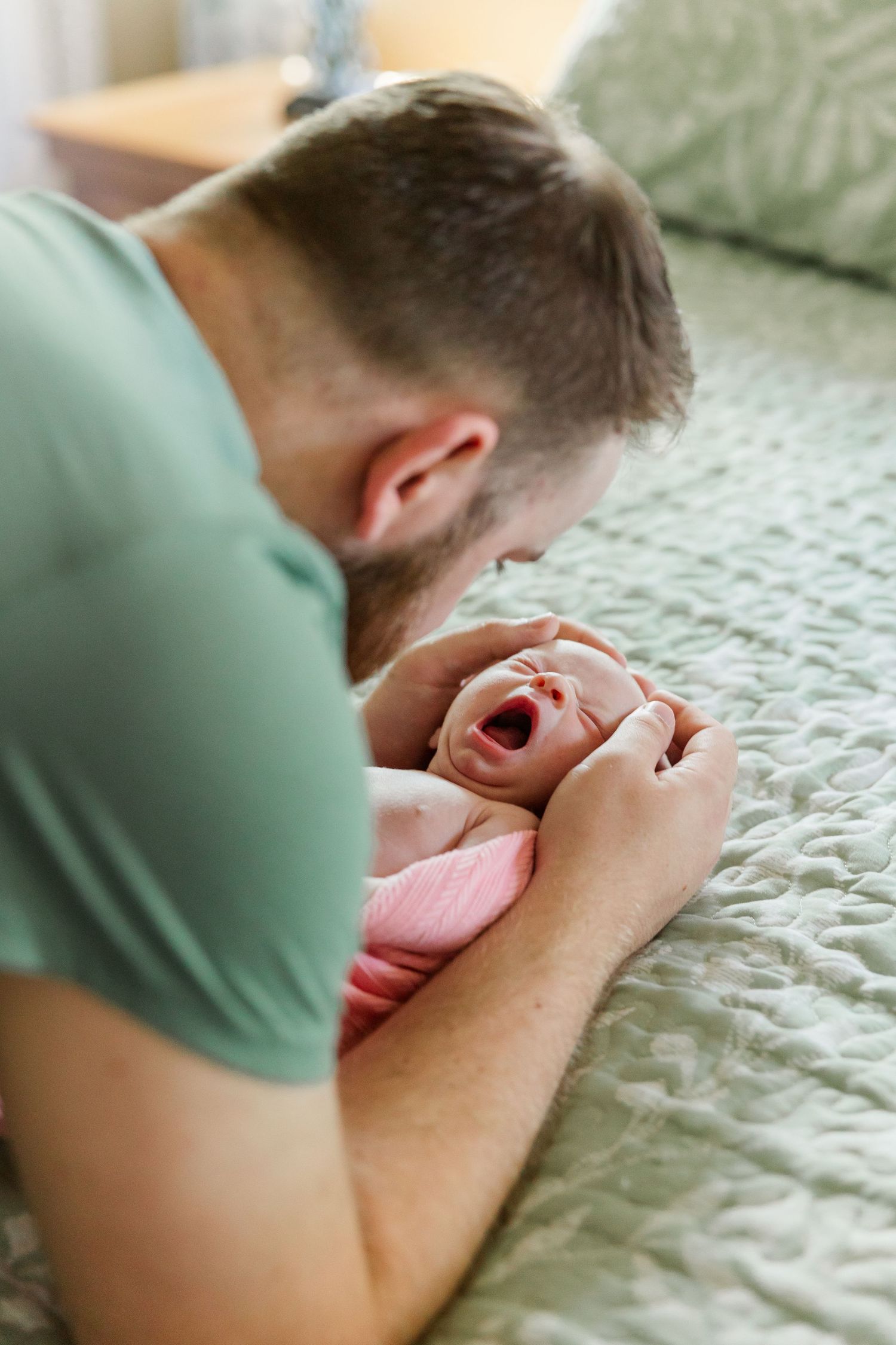 A father leans over his yawning newborn baby dressed in pink lying on a green quilted bed.