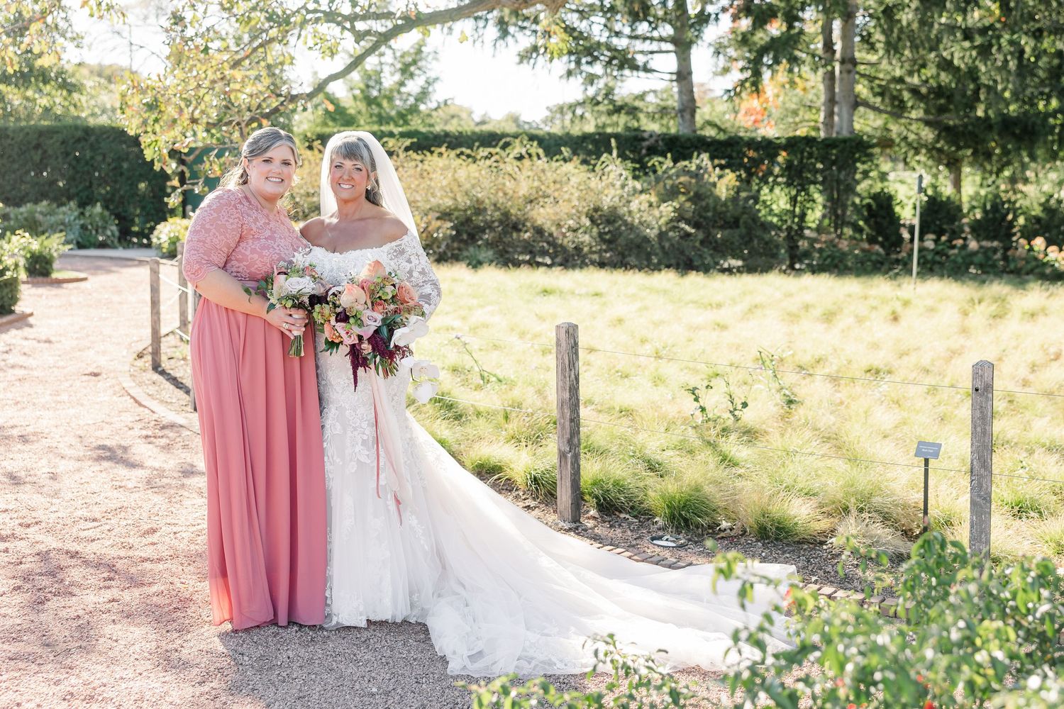 Bride in a white lace wedding gown with a long veil poses with a bridesmaid in a coral dress, both holding bouquets in a sunny garden.