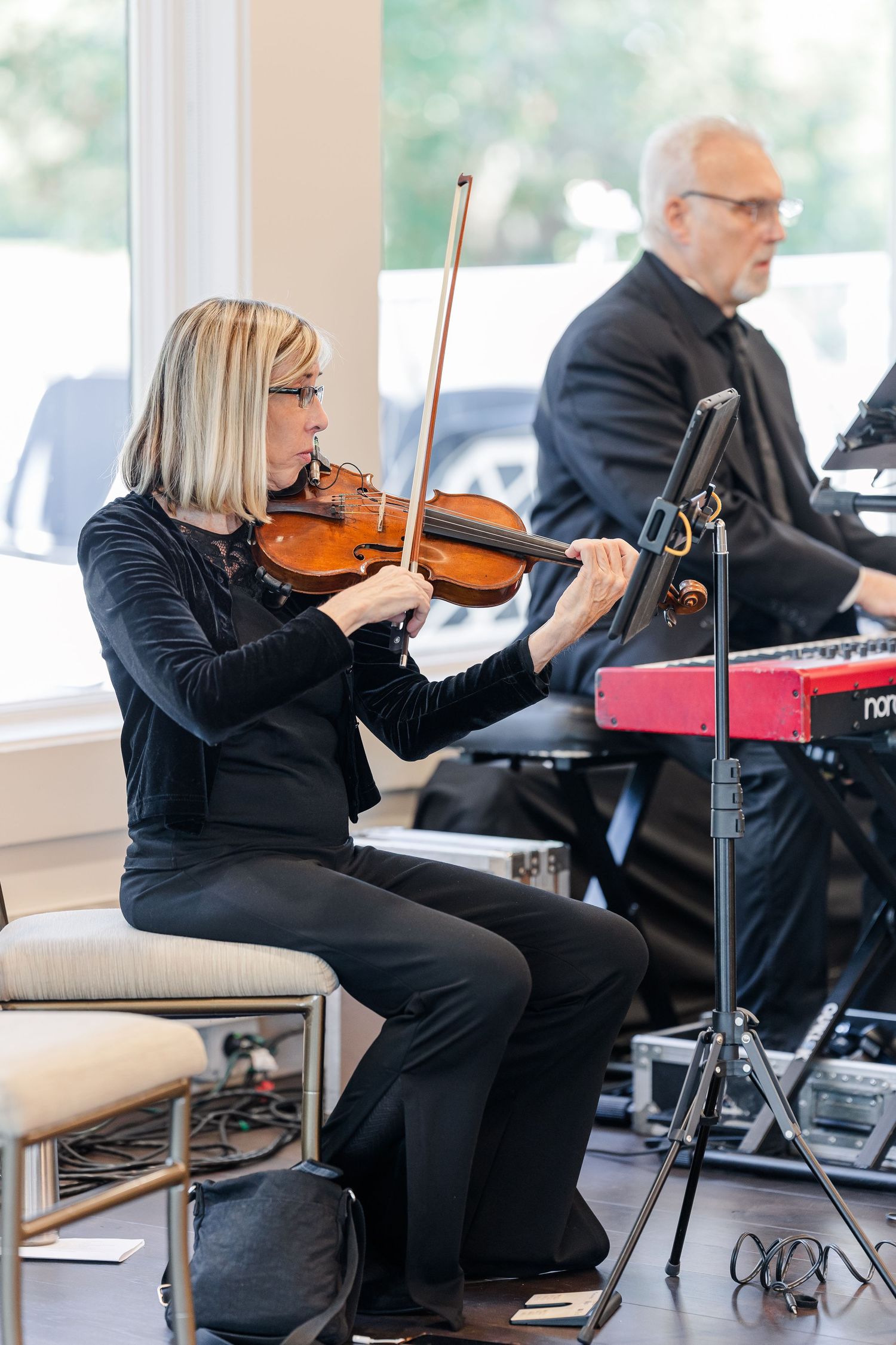 A woman plays the violin beside a music stand while a man in a dark suit plays a red keyboard at a live event near bright windows.