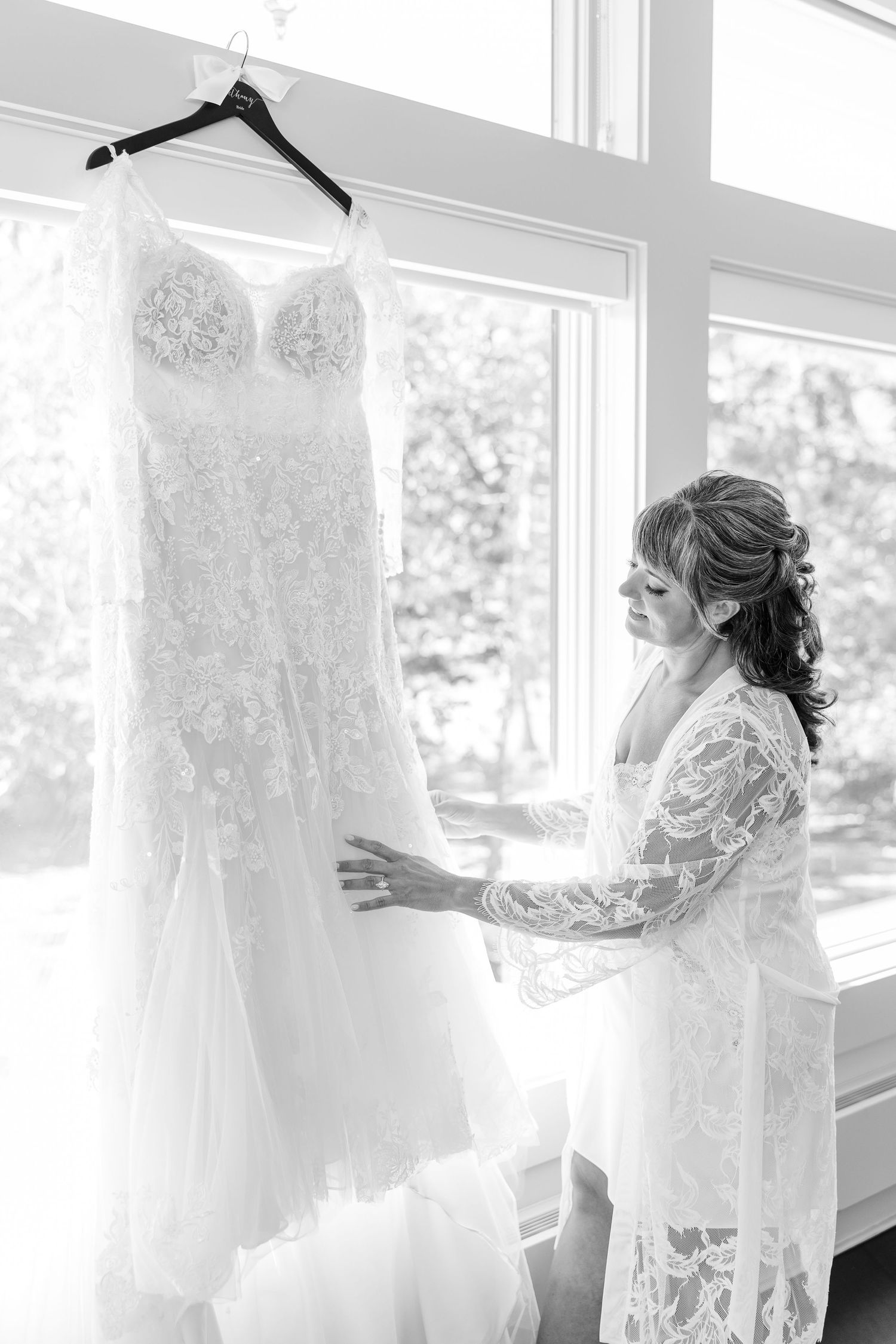 Black and white photo of a bride in a lace robe touching her lace wedding dress hanging by a bright window.