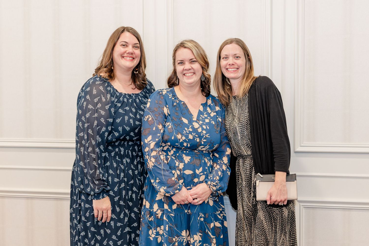 Three smiling women in floral and patterned dresses pose together at an indoor event in front of a white paneled wall.