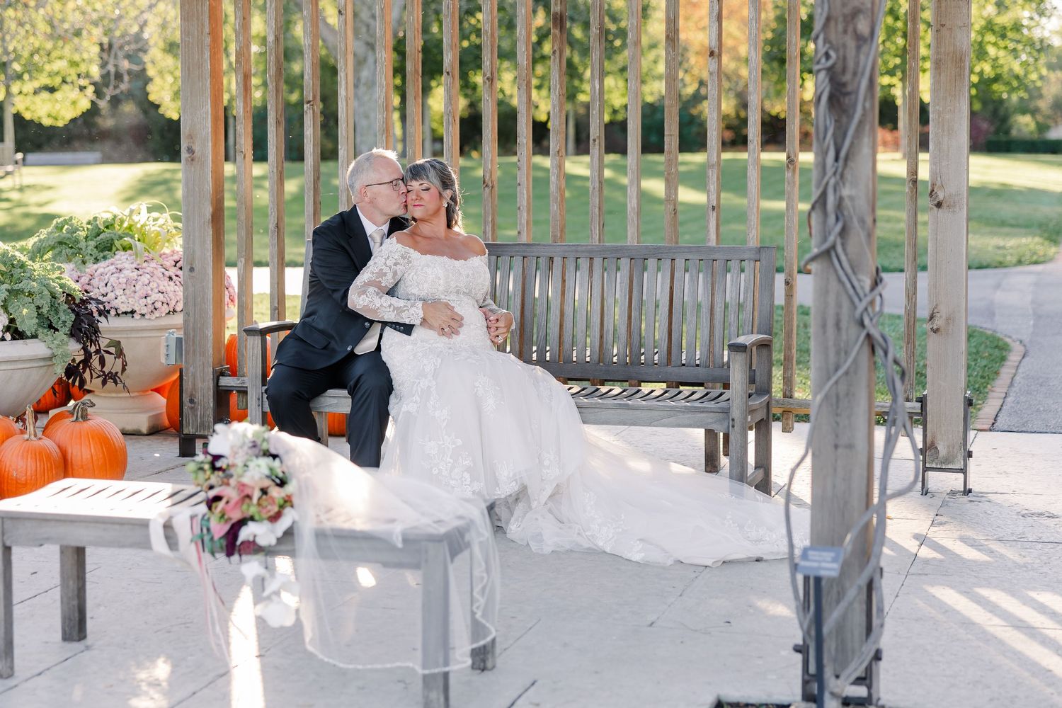 Bride in a white off-the-shoulder gown and groom in a dark suit share an embrace on a wooden garden bench beside autumn pumpkins.