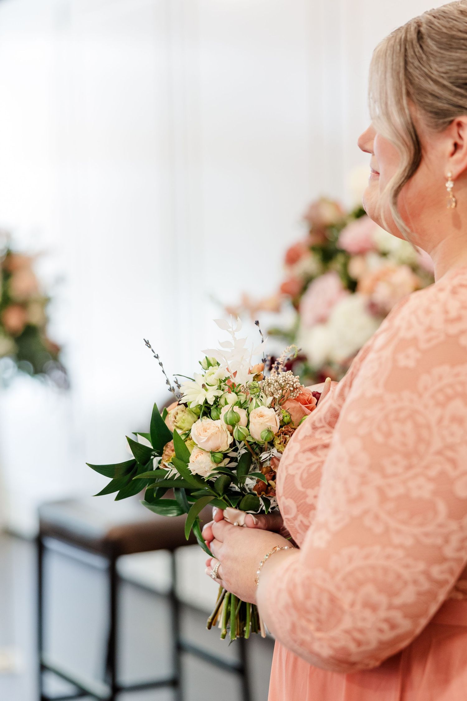 Woman in a coral lace dress holds a bouquet of blush roses and greenery at a wedding, with floral arrangements softly blurred behind her.