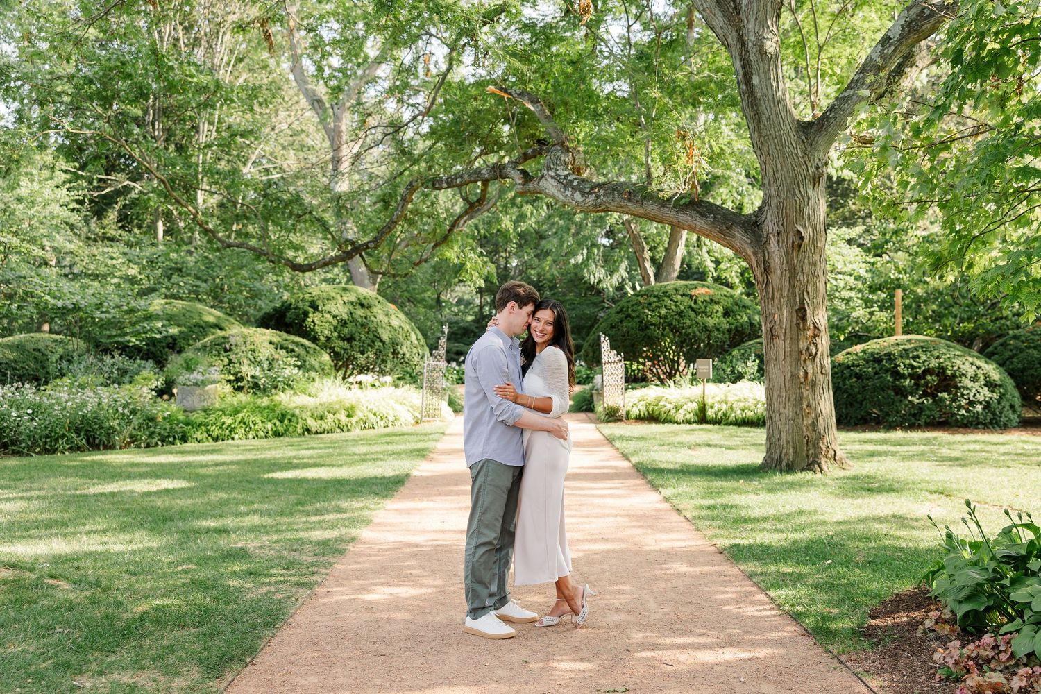 Newly engaged Alysia and Jack kiss beneath the trees along a garden pathway.