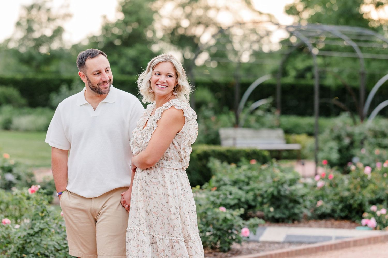 Smiling couple walking together in a rose garden at golden hour, woman in floral dress.