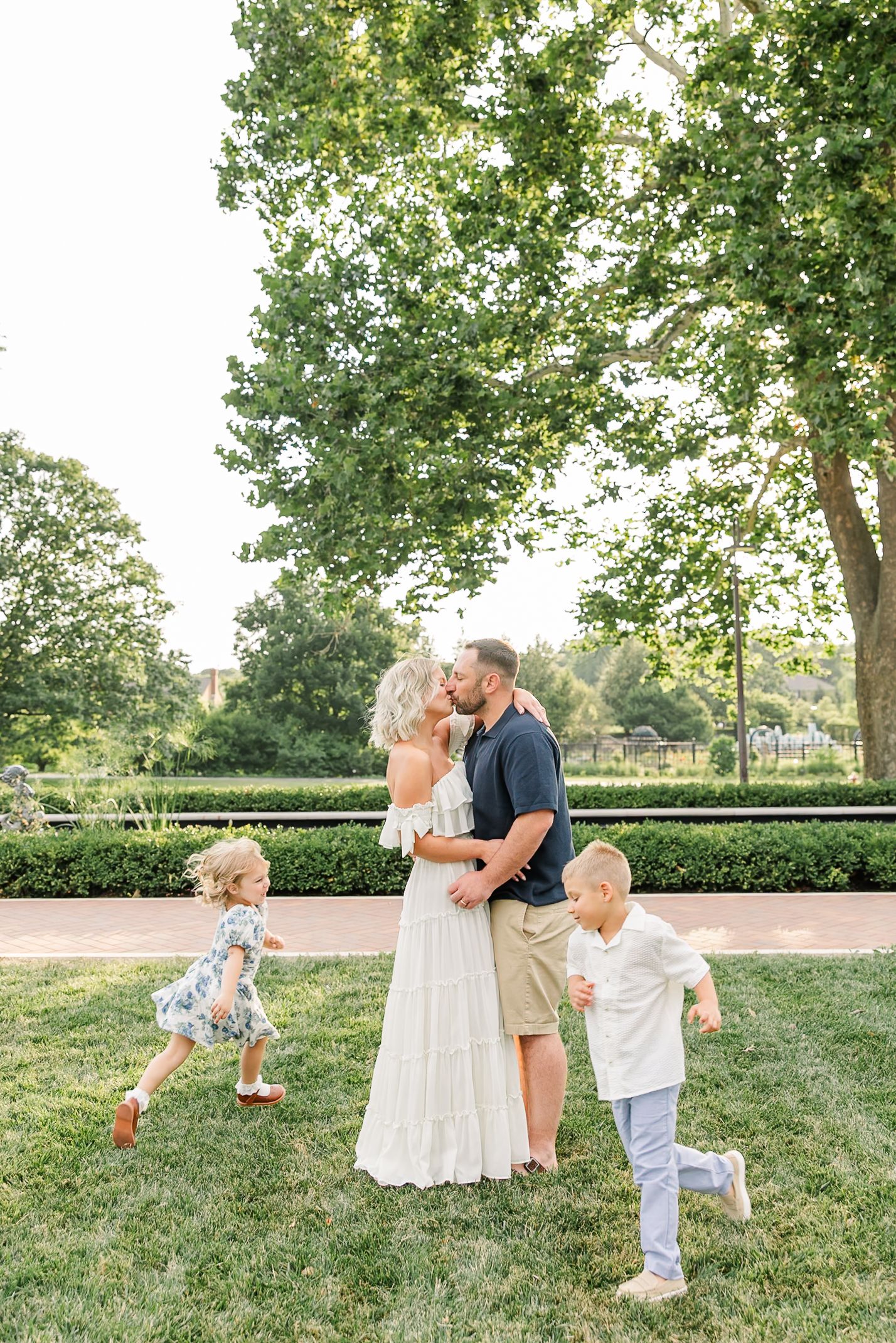 A couple shares a kiss while two young children play around them in a lush green park during a family photo session.