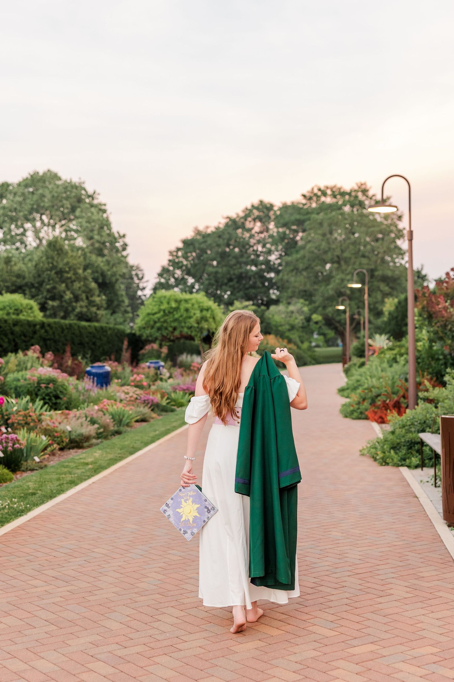 Samantha carries her cap and graduation gown along a garden path during her Bartlett High School senior portraits.