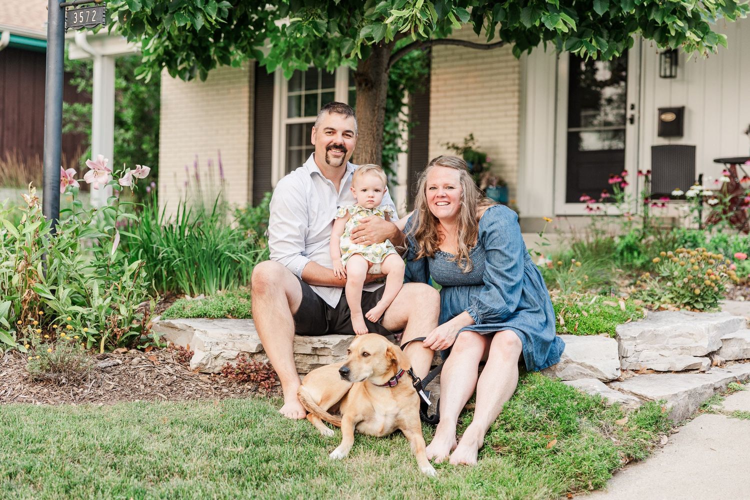 Family moment with pet dog in front of house surrounded by greenery and flowers.
