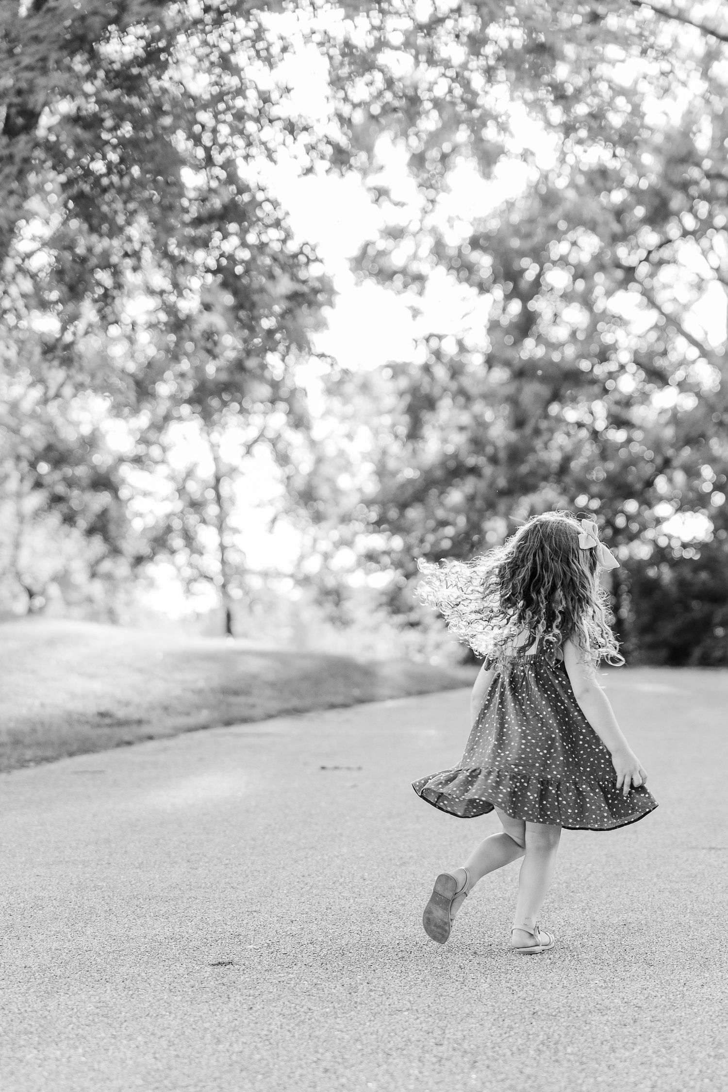 Black and white photo of child twirling in dress on sunny park path.