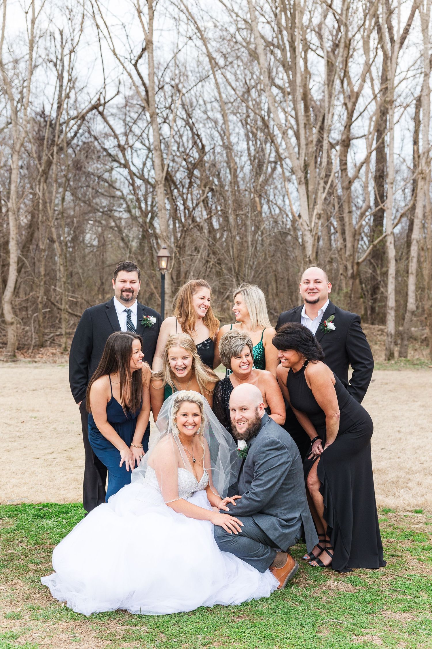 Wedding party gathers for a fun group photo in a wooded outdoor setting.