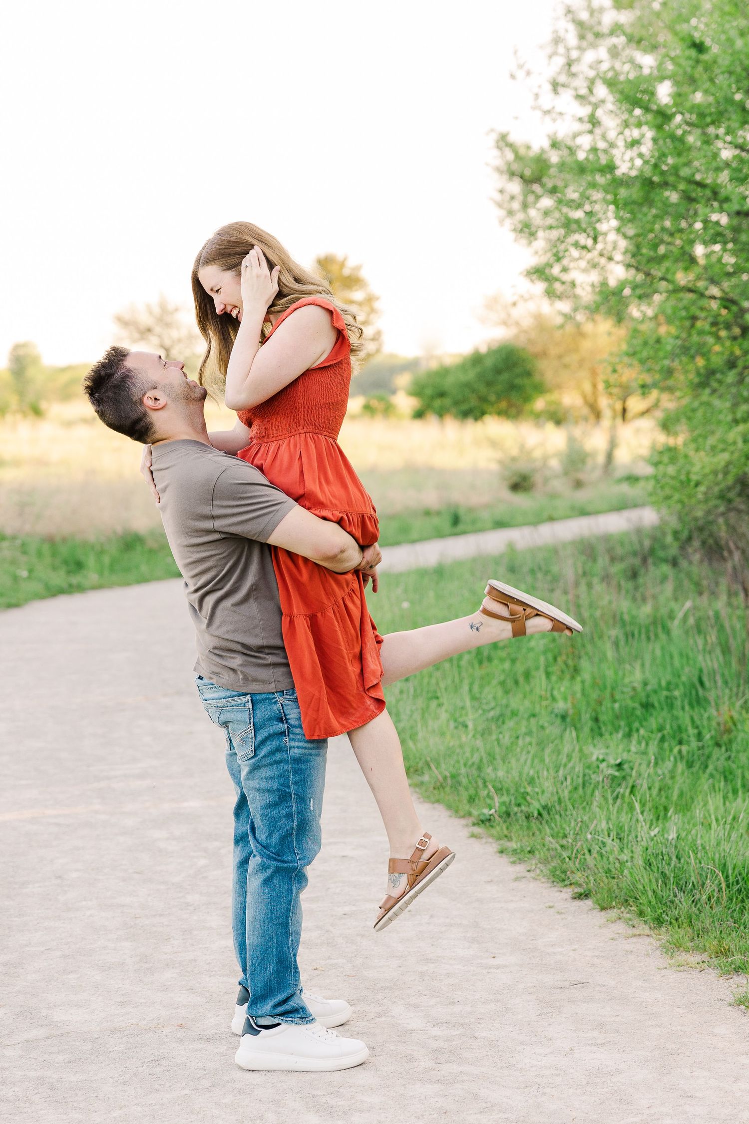 A couple shares a romantic moment on a dirt path as he lifts her up in her red dress against a nature backdrop.