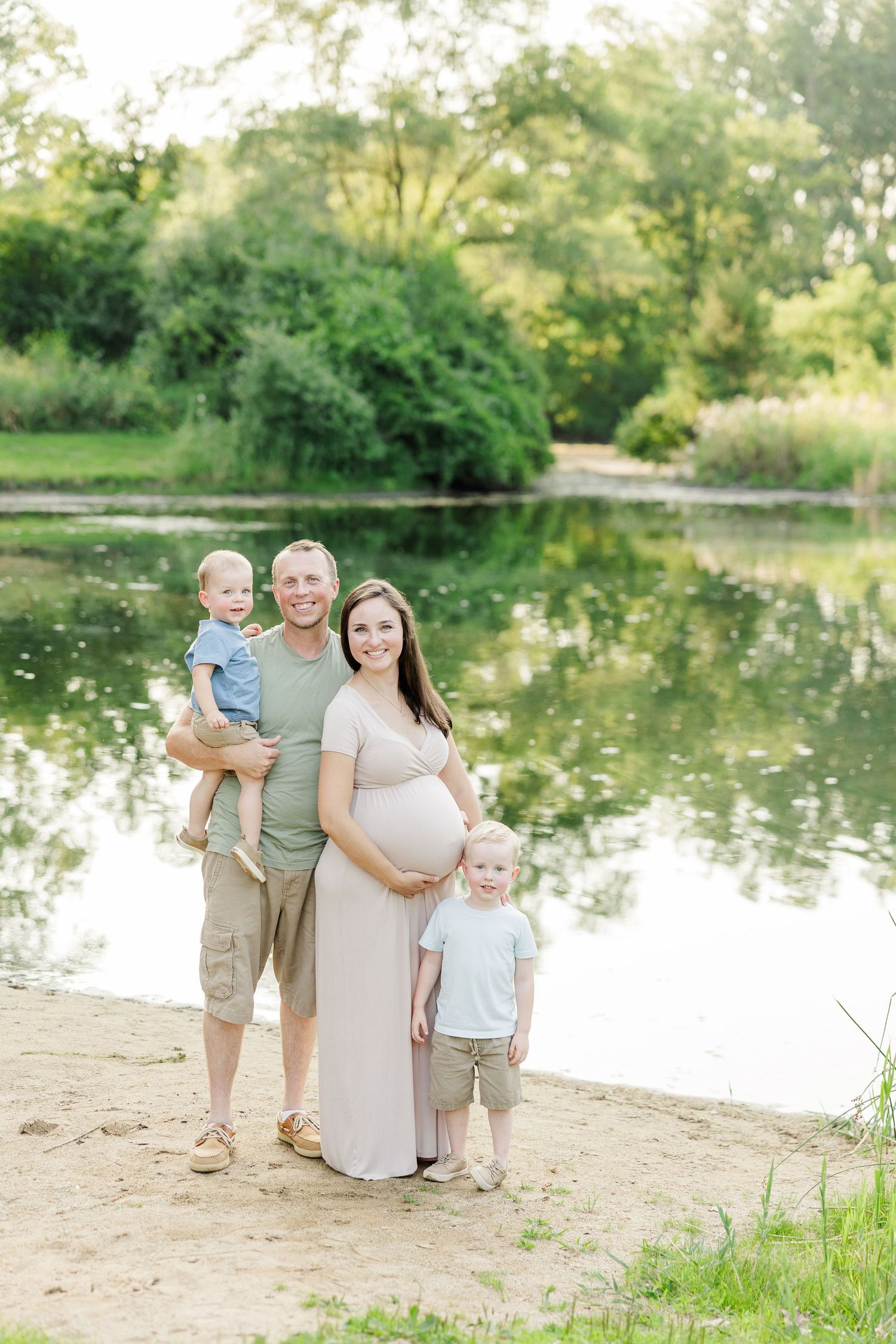 A family stands together near a serene pond surrounded by lush greenery on a sunny summer afternoon.