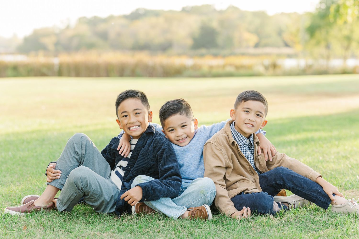 Three young boys sit together on grass in a park on a sunny day wearing casual clothes. Busse Woods