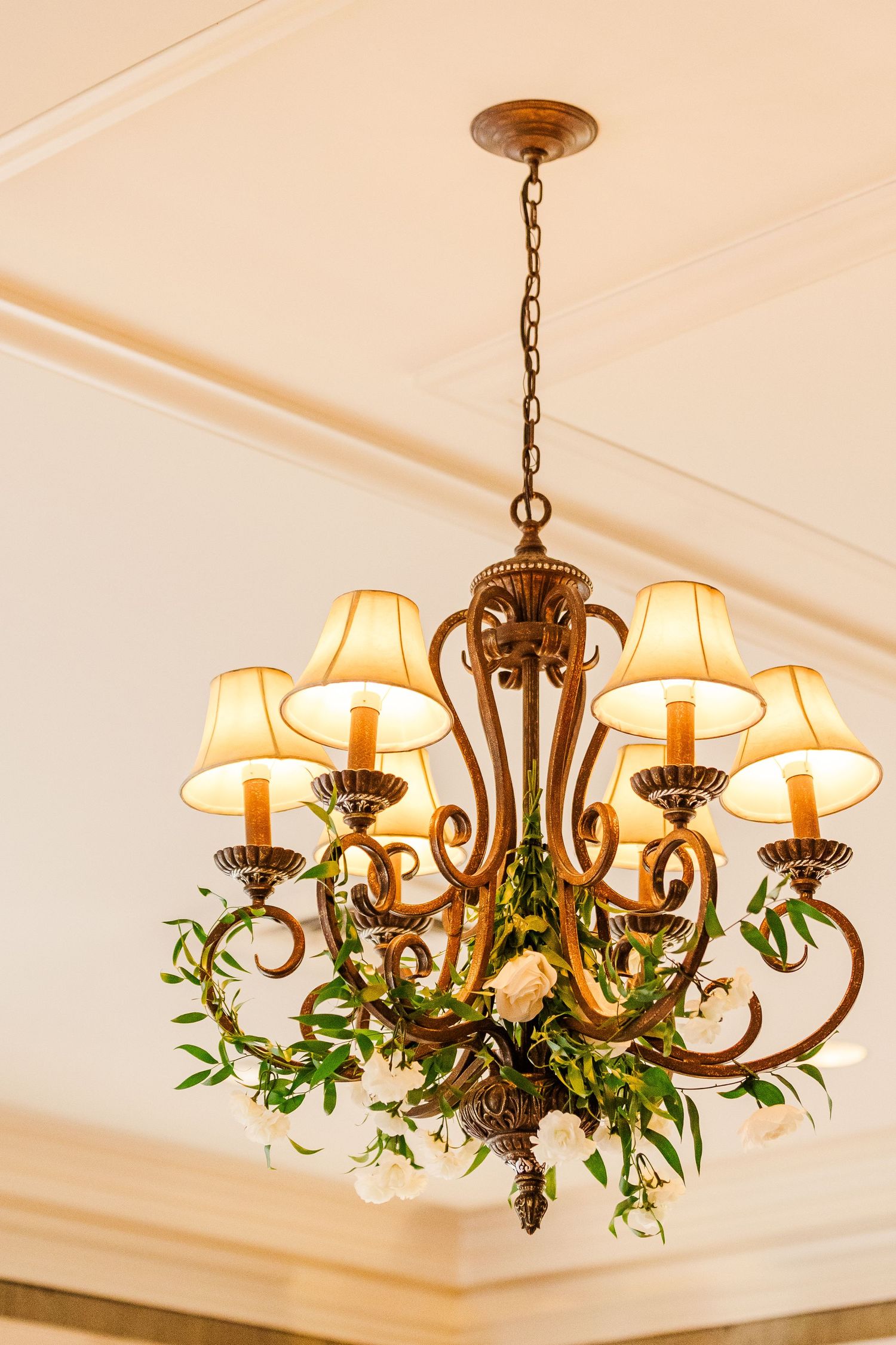 Ornate brass chandelier with cream lampshades decorated with fresh greenery for wedding decor.