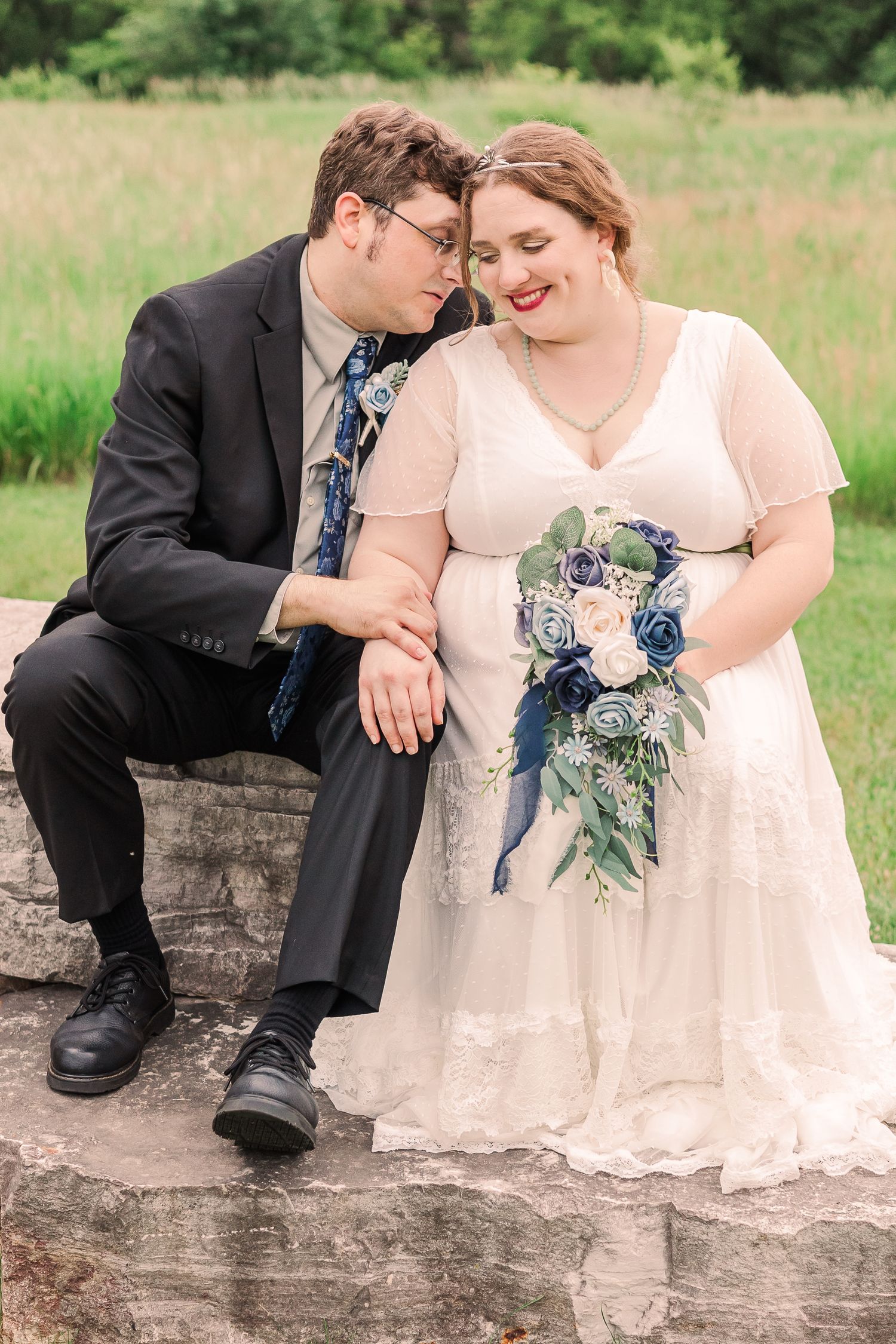 A tender wedding moment captured as the couple sits together on a stone wall in a meadow.