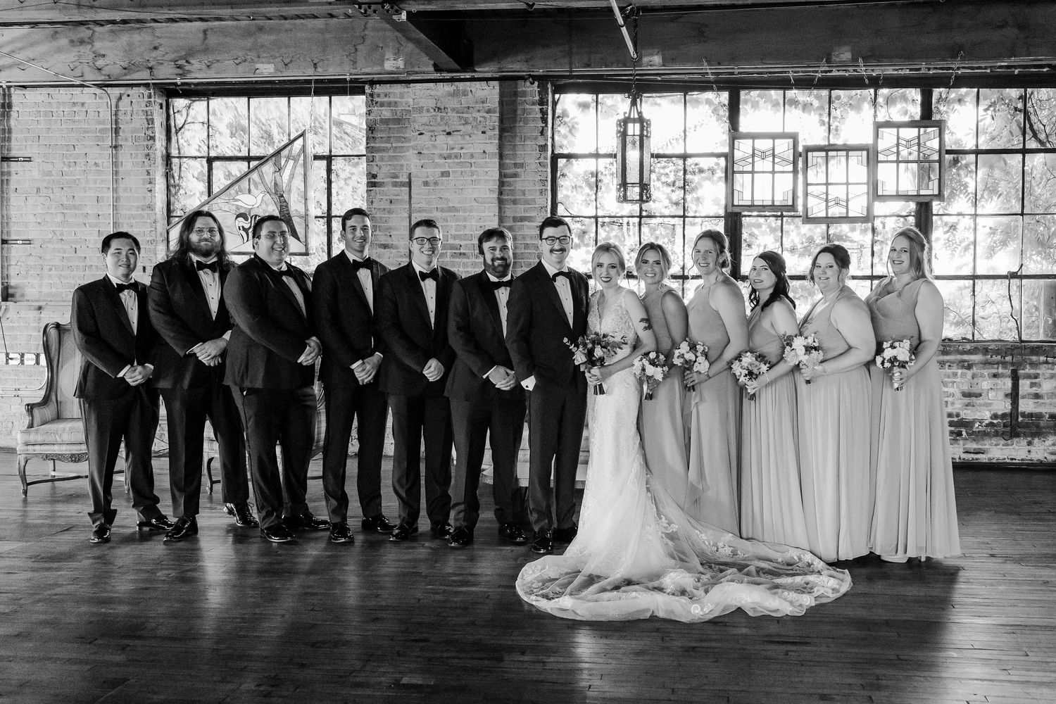 Black and white photo of wedding party lined up in formal attire against industrial brick backdrop.