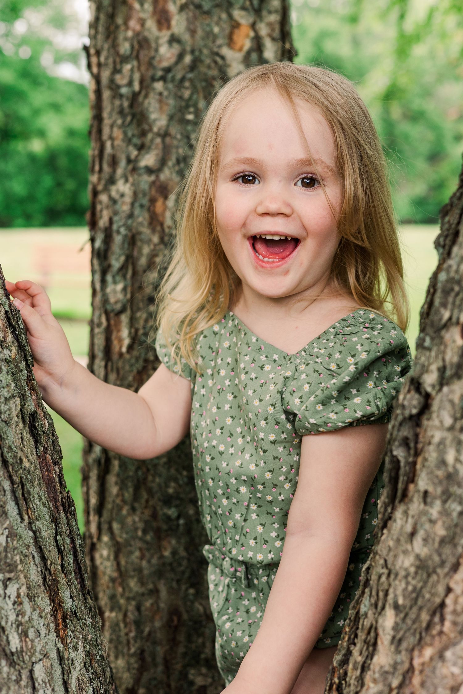 A cheerful young child in a green floral dress poses playfully between two tree trunks outdoors.
