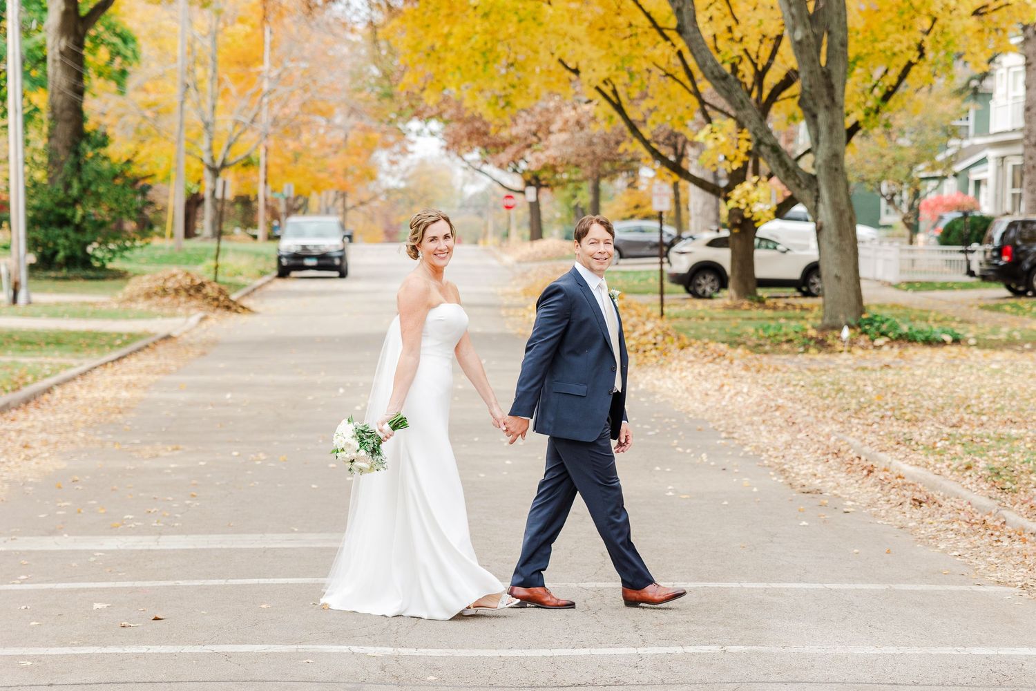 Wedding couple holding hands while walking on street with yellow fall trees.