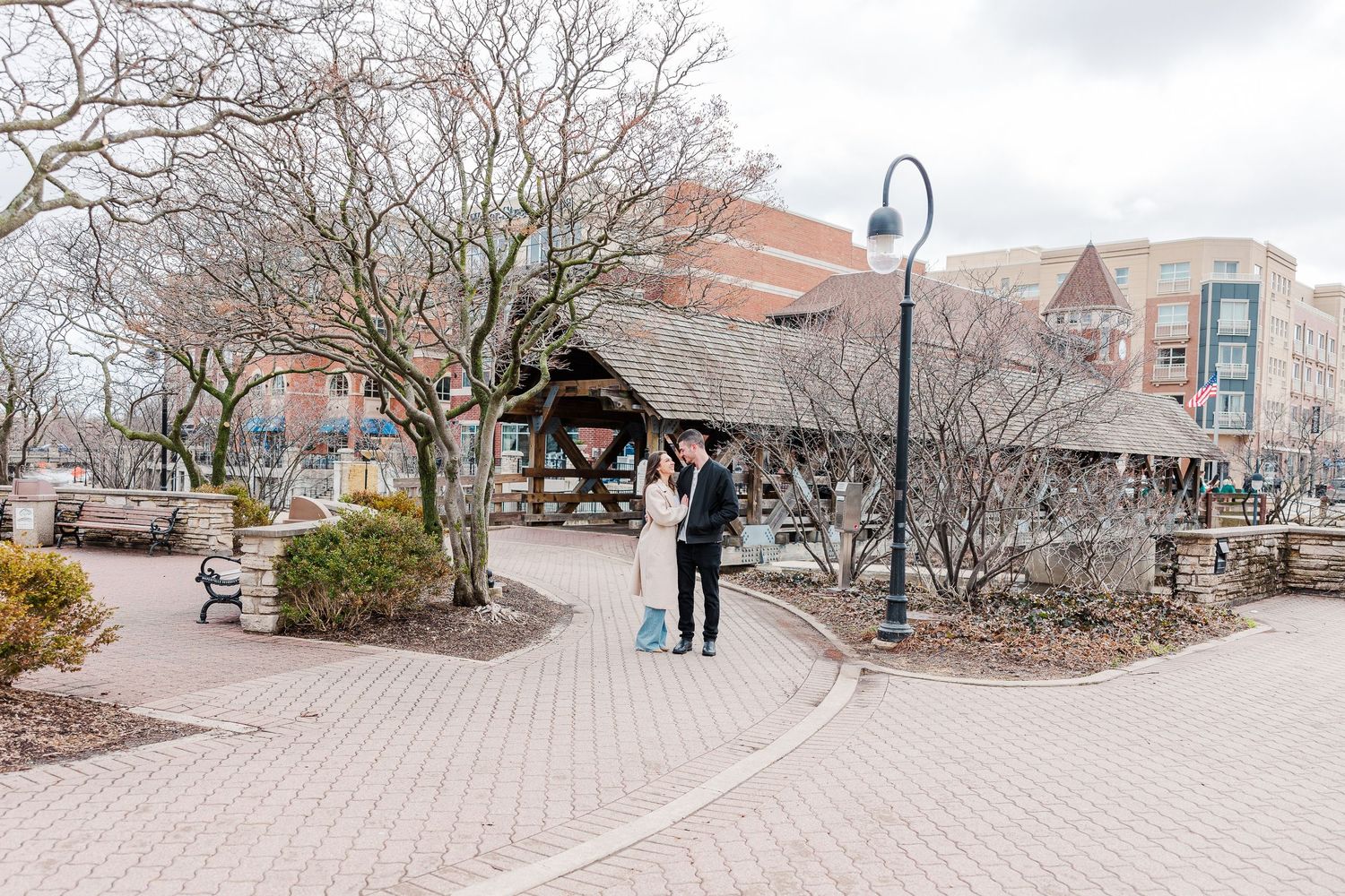 A couple stands together on a curved brick walkway near a wooden pavilion structure.