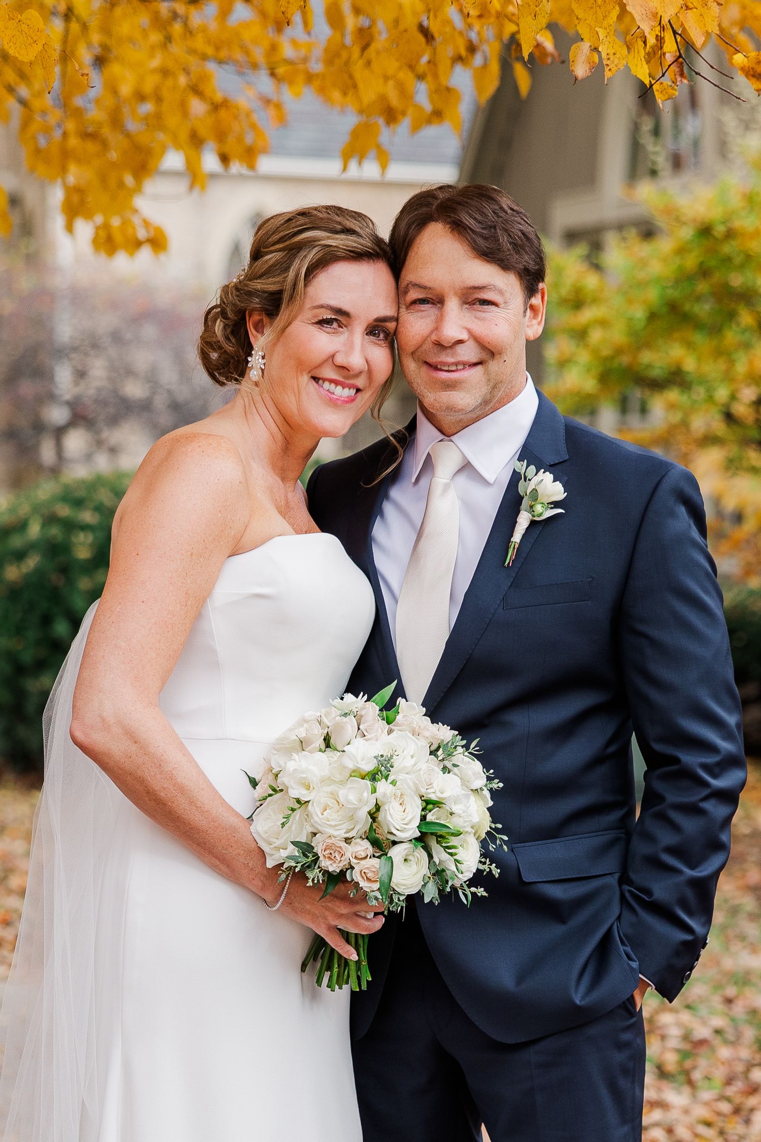 Wedding couple embracing outdoors in autumn with golden leaves and white bouquet.