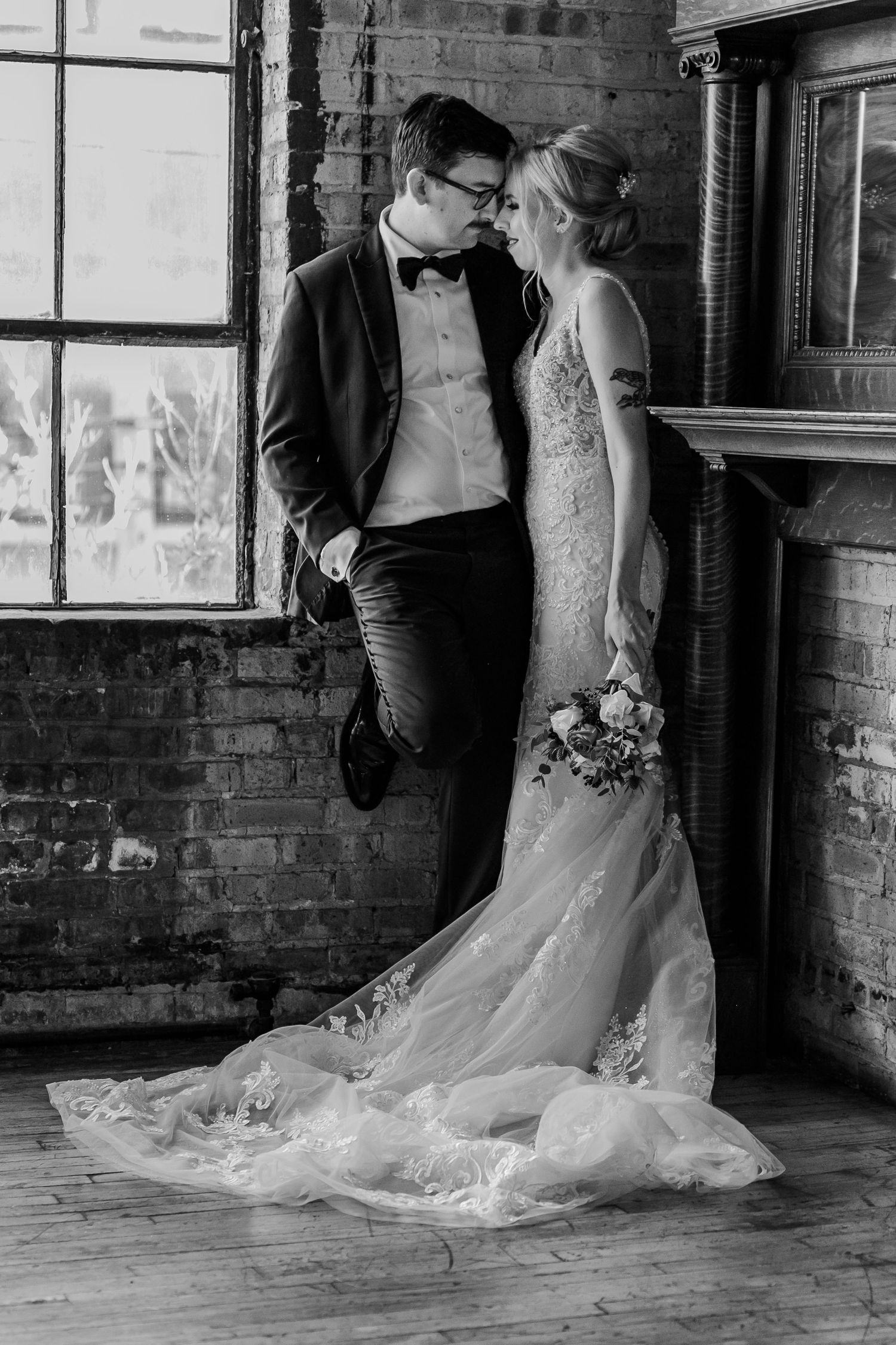 Elegant wedding couple poses against exposed brick wall with dramatic window lighting.