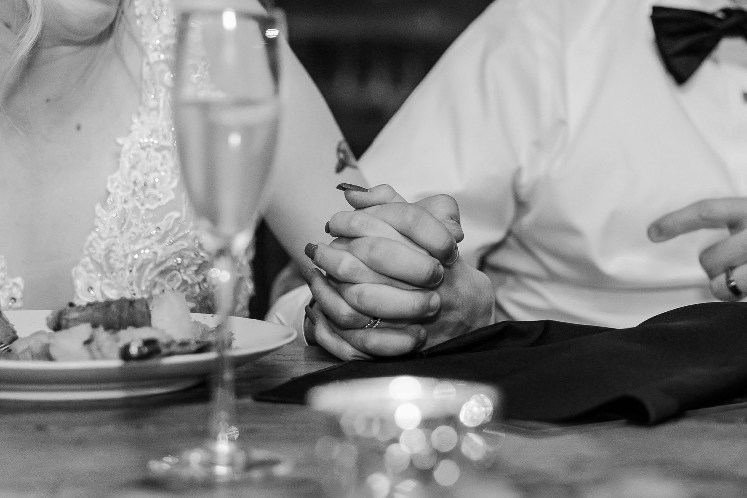 Black and white close-up of clasped hands at wedding reception table with champagne glass.