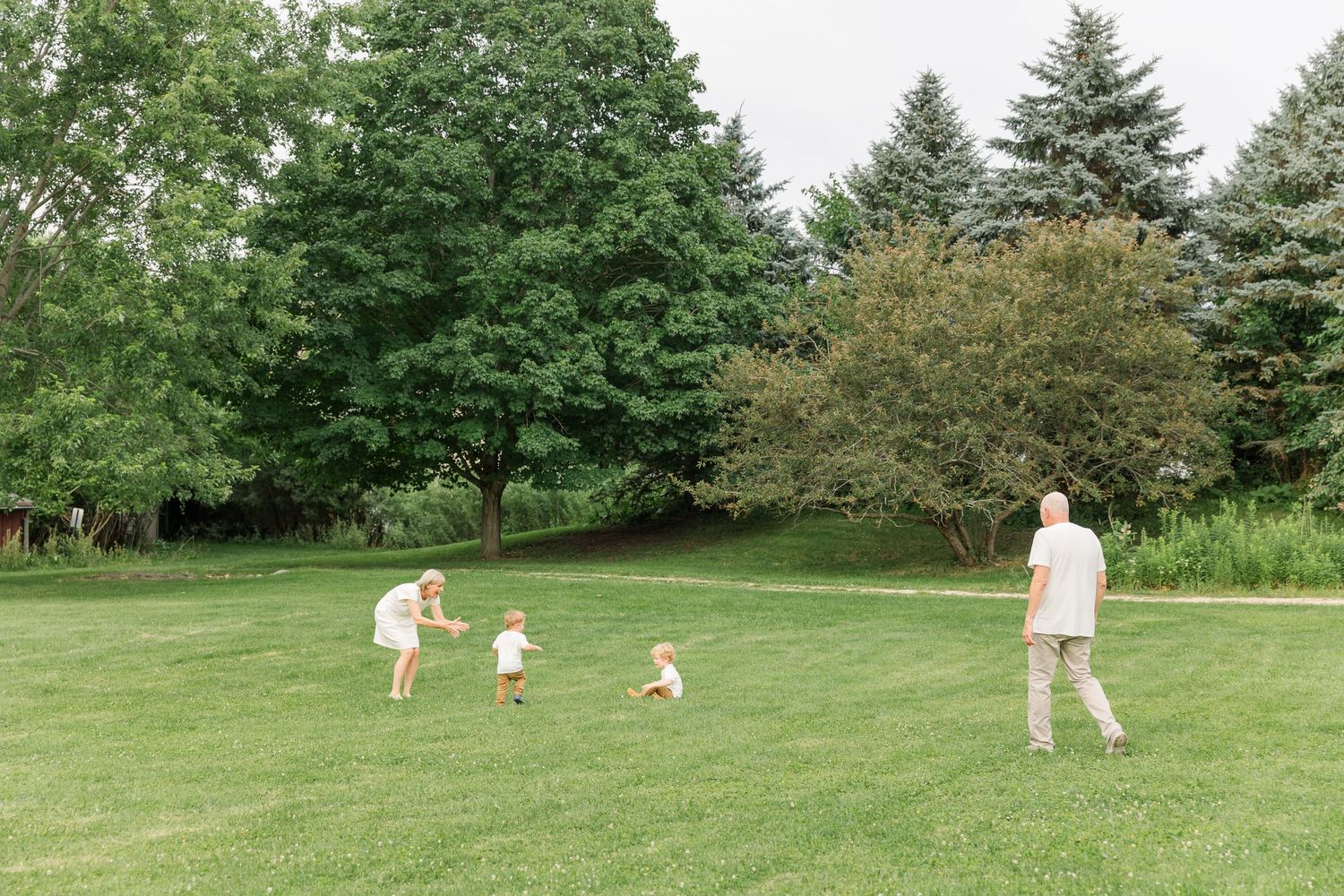Family playing together on lush green lawn surrounded by trees with children running toward mother.