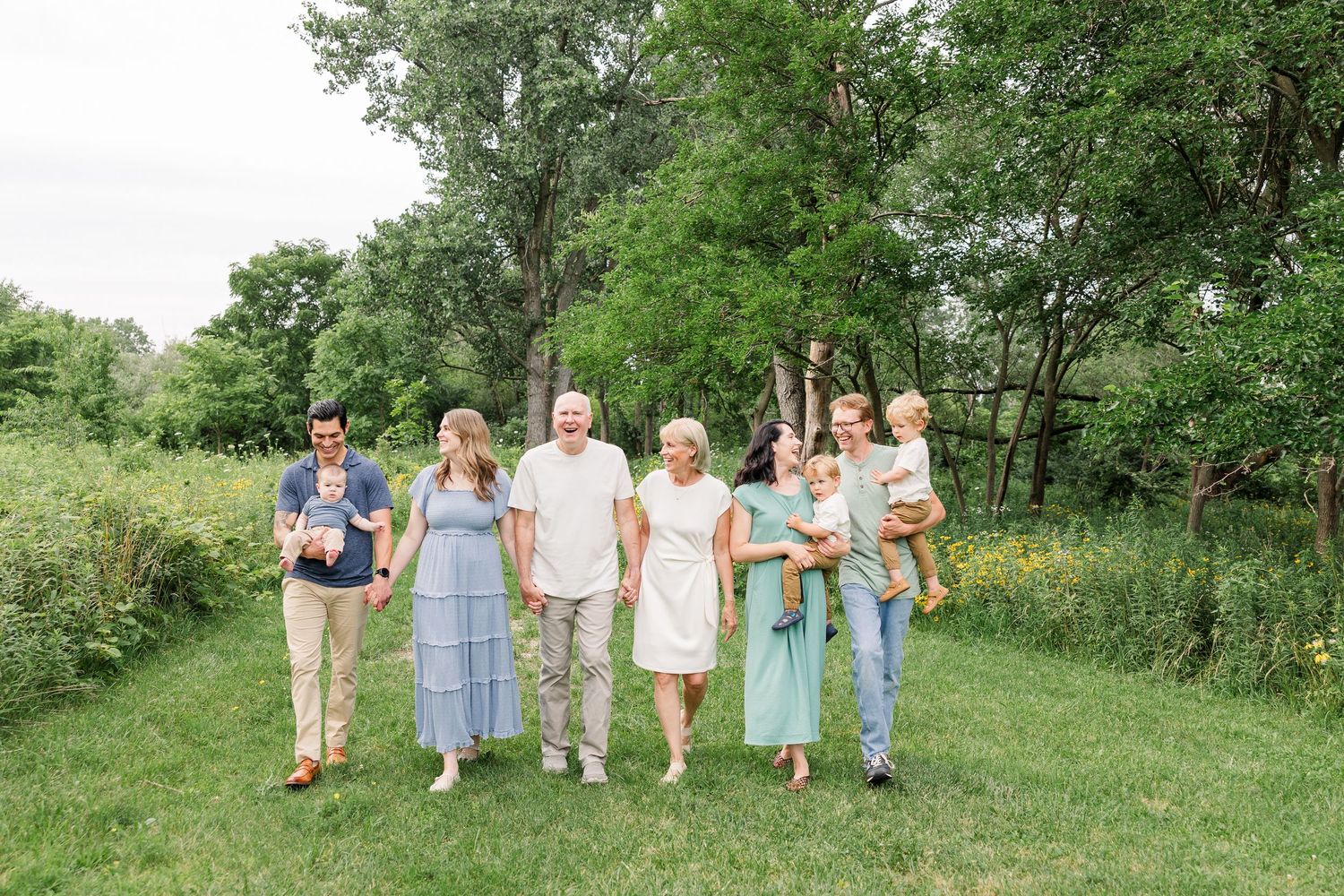 A multigenerational family walks together on green grass surrounded by lush trees in coordinated blue and neutral outfits.