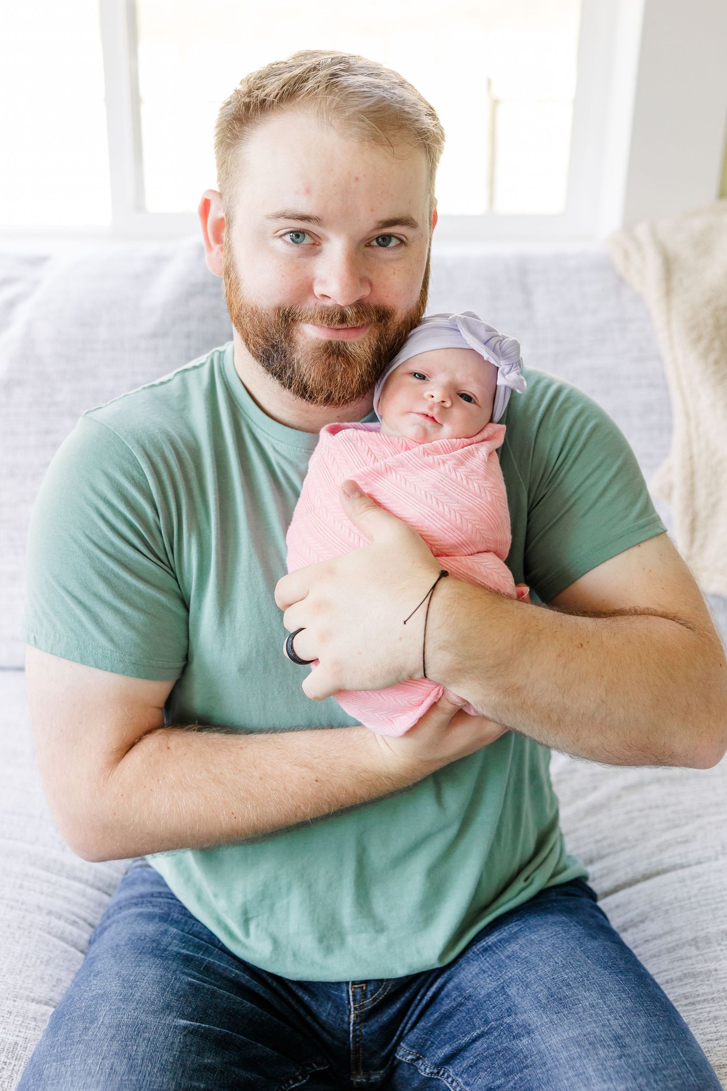 Bearded father in green t-shirt smiling while holding swaddled newborn baby girl in pink wrap and floral headband.