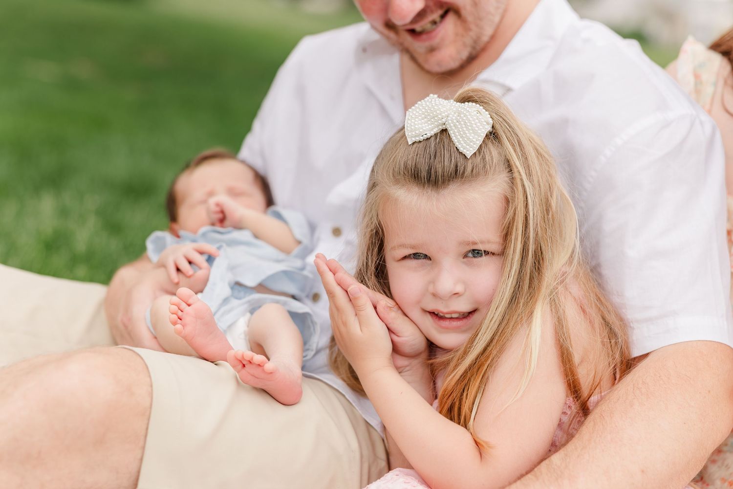 A smiling blonde girl with a bow poses beside her father holding a newborn baby on a blanket outside.
