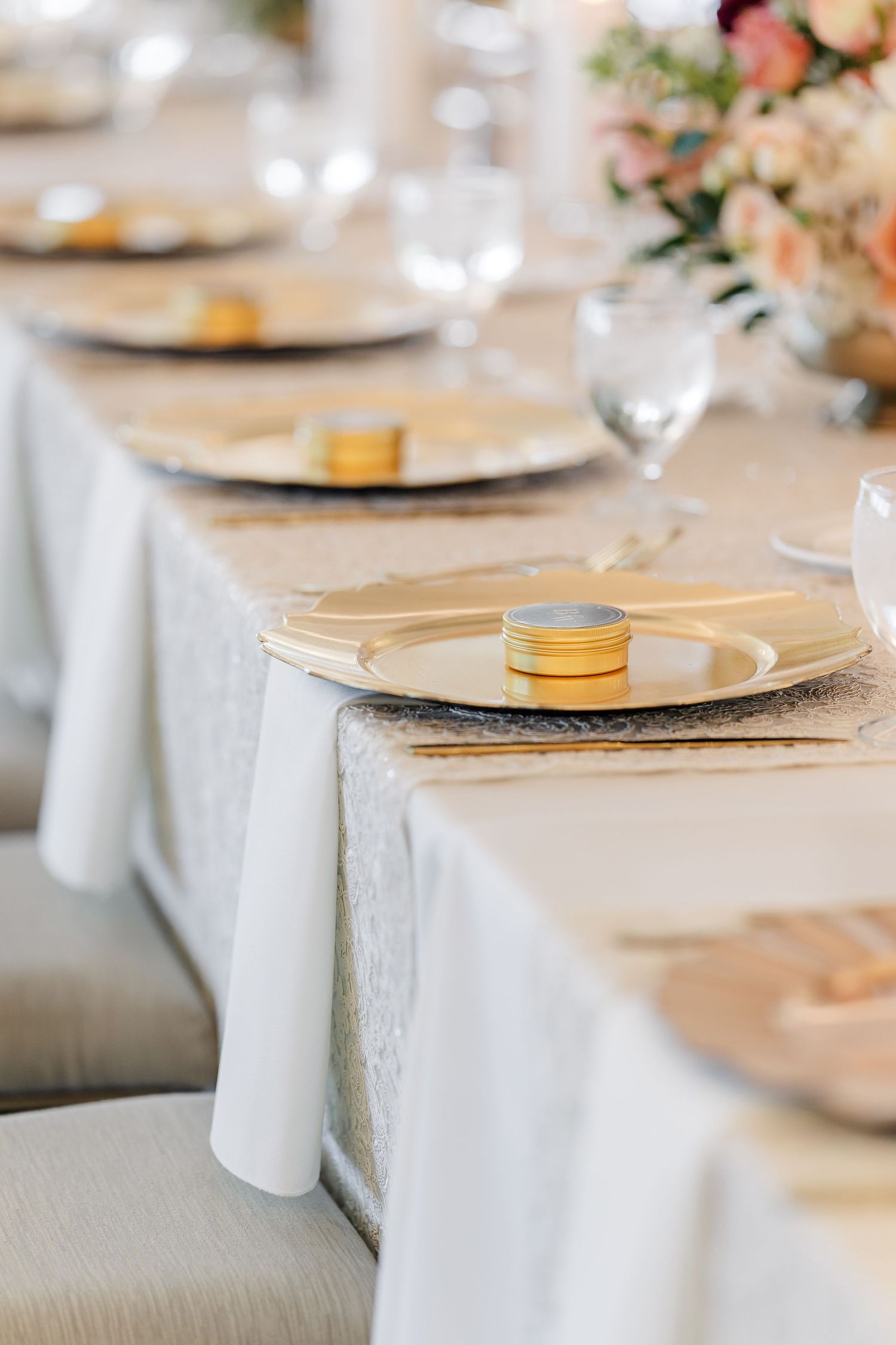 Elegant wedding reception table with gold charger plates, small gold tins, white linens, wine glasses and a peach floral centerpiece.