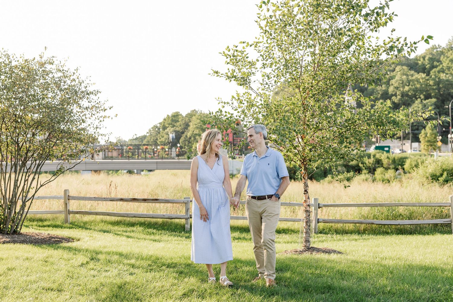 Couple holding hands and smiling at each other while walking on green grass near a wooden fence outdoors.