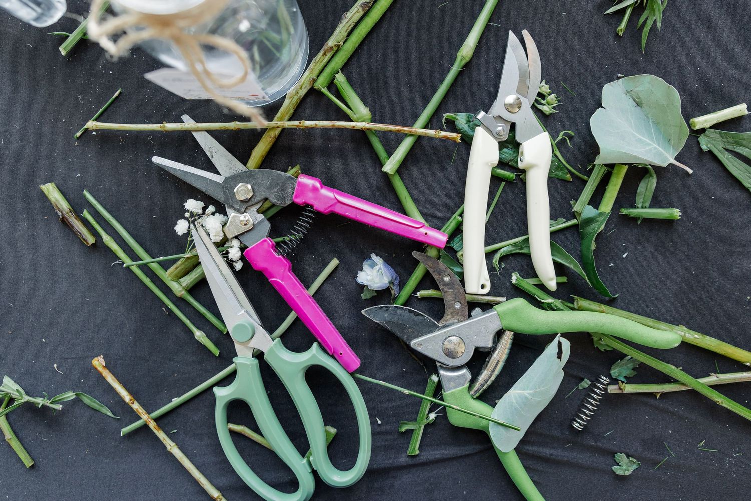 Floral arrangement tools including scissors and pruning shears scattered among cut green stems on a dark surface.