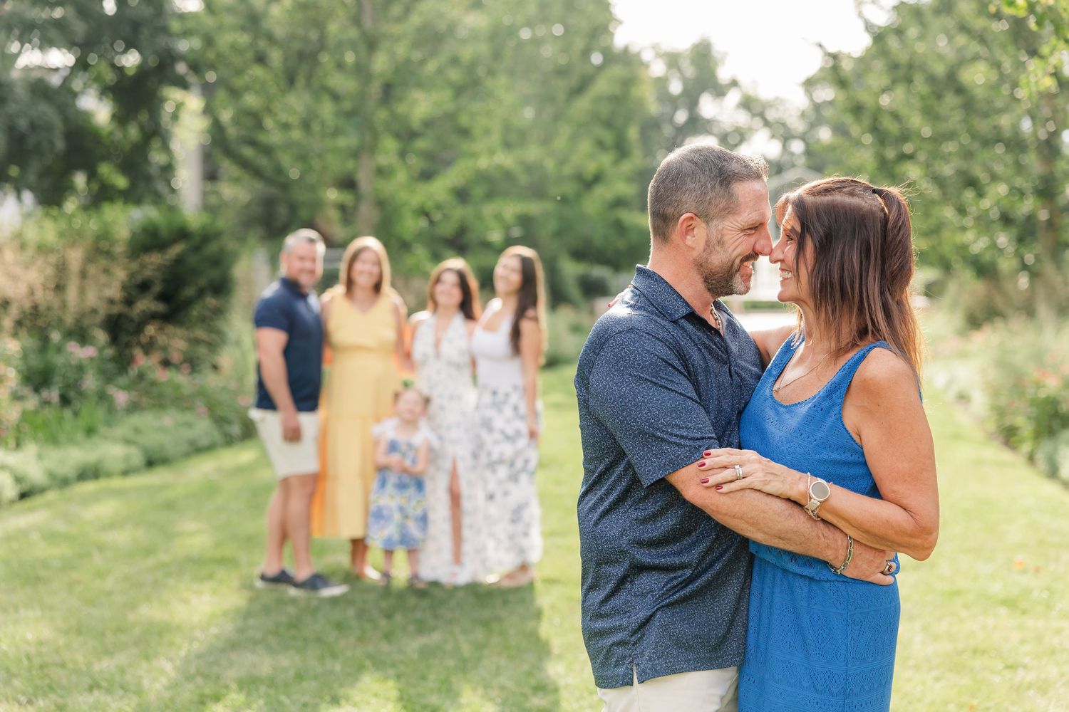 Couple sharing romantic moment in garden with family members standing softly blurred in background during outdoor photo session.