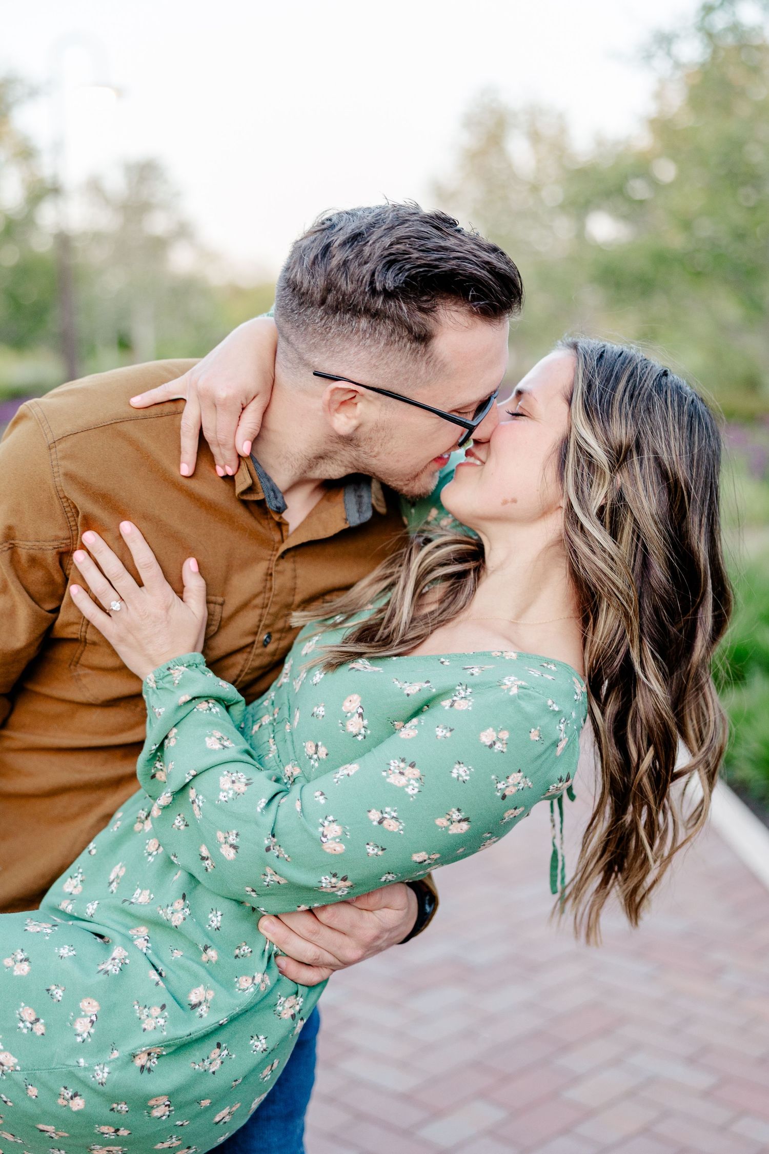 A couple shares a romantic kiss outdoors, with the woman in a green floral top and man in a brown shirt embracing.