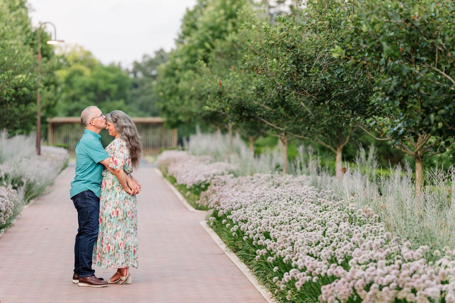 Couple embracing on a garden path surrounded by blooming lavender flowers and lush green trees at dusk.