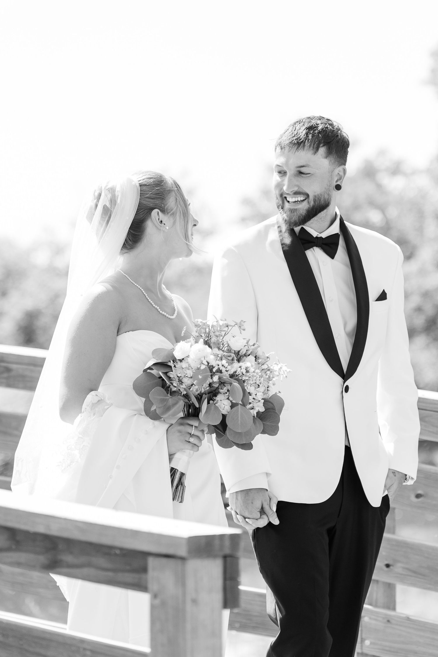 Black and white photo of smiling groom in white tuxedo holding hands with bride carrying bouquet on wooden bridge.